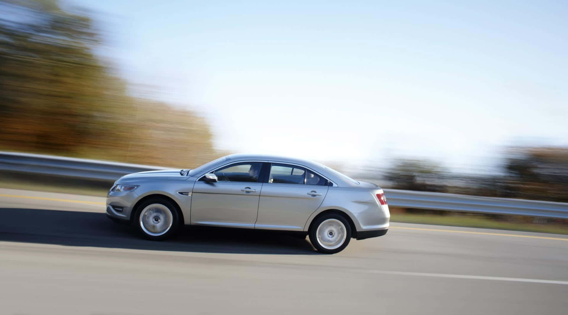 Ford Taurus 桌布 壁紙 — 一輛銀色轎車在高速公路上疾馳，背景是模糊的秋樹和護欄。