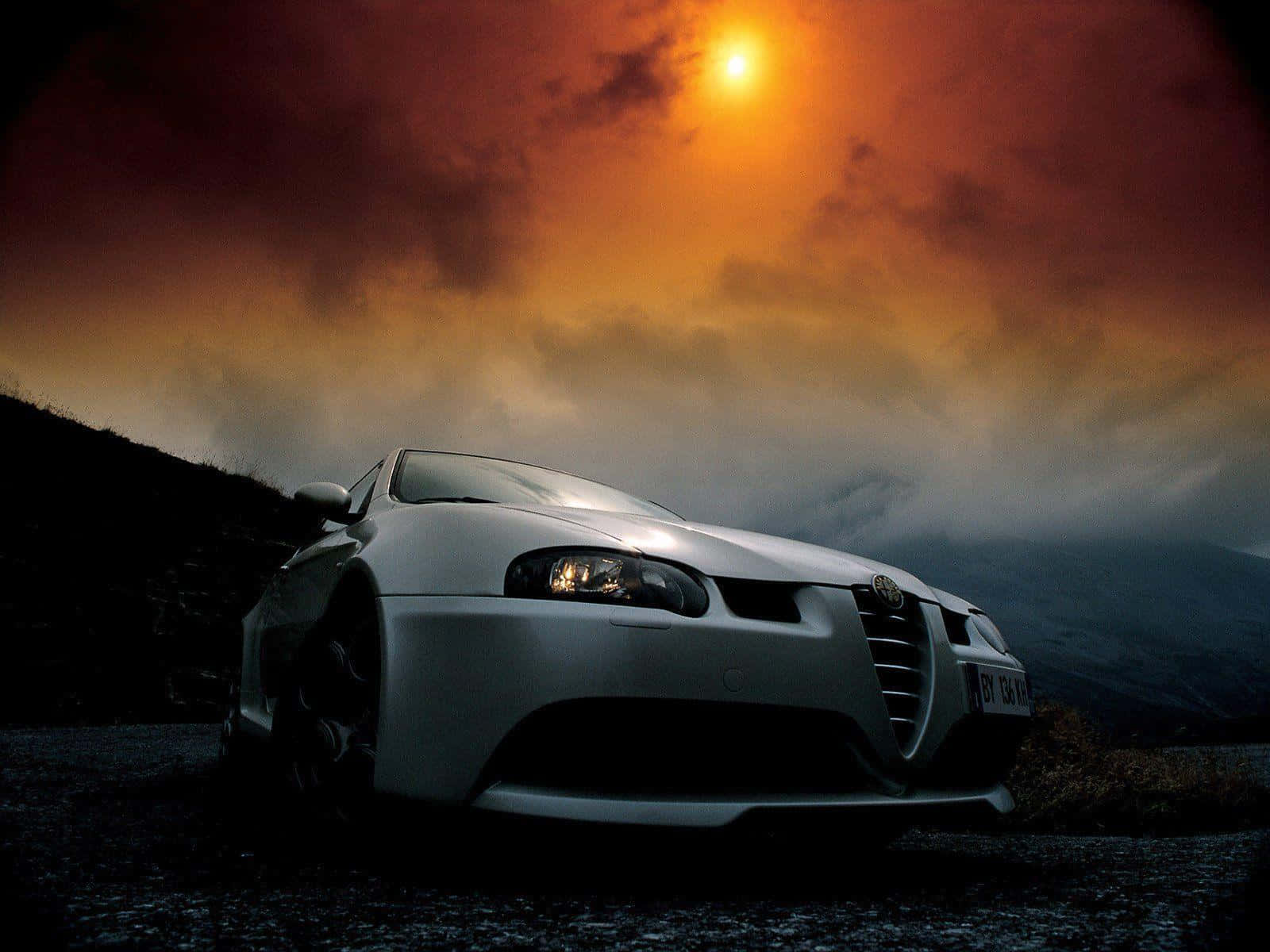 Alfa Romeo Gt 桌布 壁紙 — 一輛銀色跑車在火紅的日落天空下閃耀，伴隨著戲劇性的雲彩和遠山。