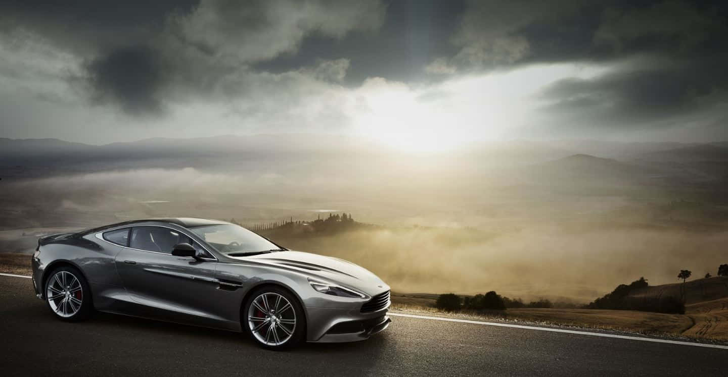 Aston Martin Dbs Superleggera 桌布 壁紙 — 一輛時尚的銀色跑車沿著蜿蜒的道路行駛，穿過霧氣瀰漫的起伏山丘，天空陰雲密布，氣氛戲劇化。