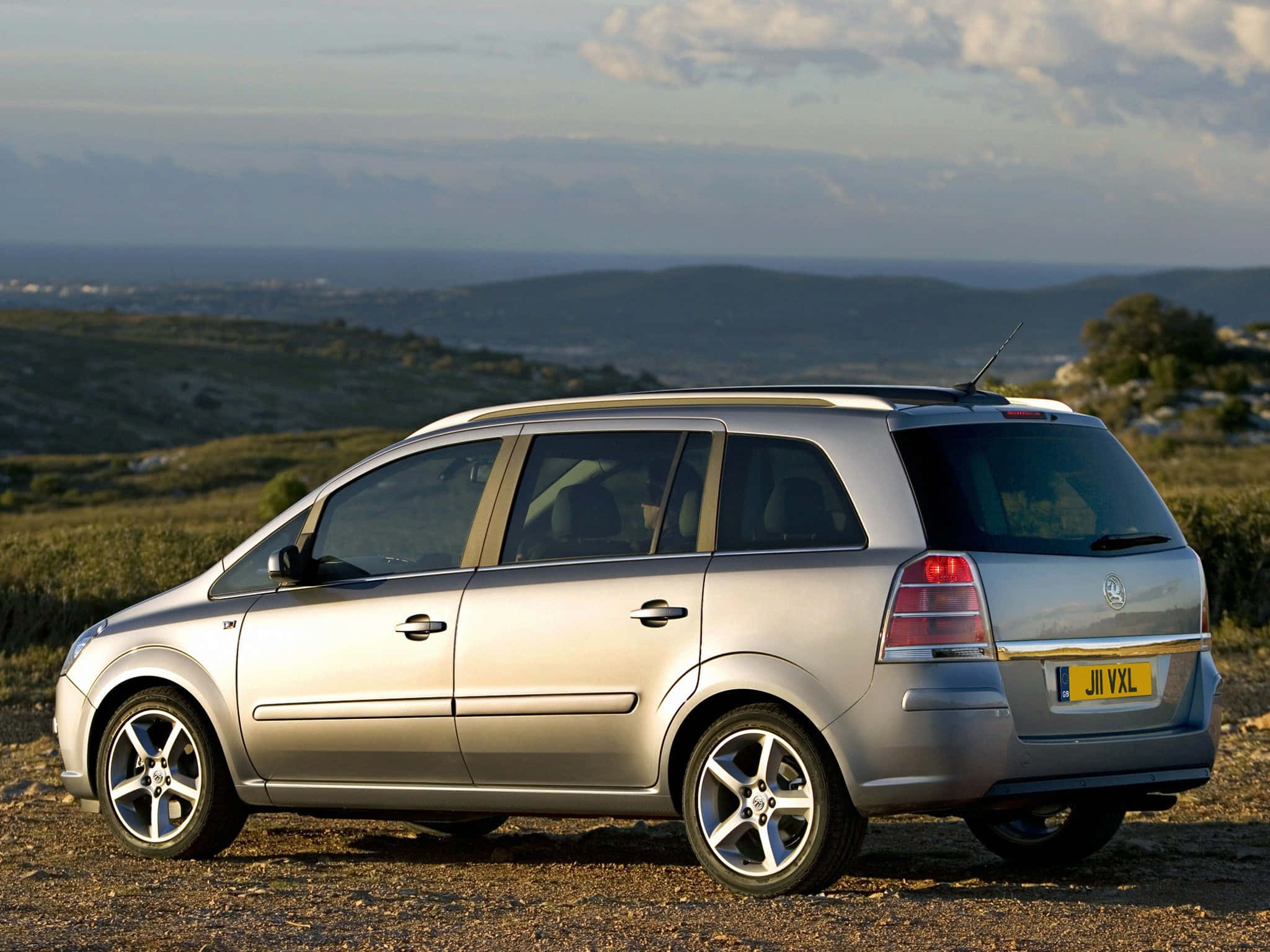 Opel Zafira 桌布 壁紙 — 一輛銀色旅行車停泊在碎石山坡上，俯瞰多雲天空下的遙遠海岸景觀。