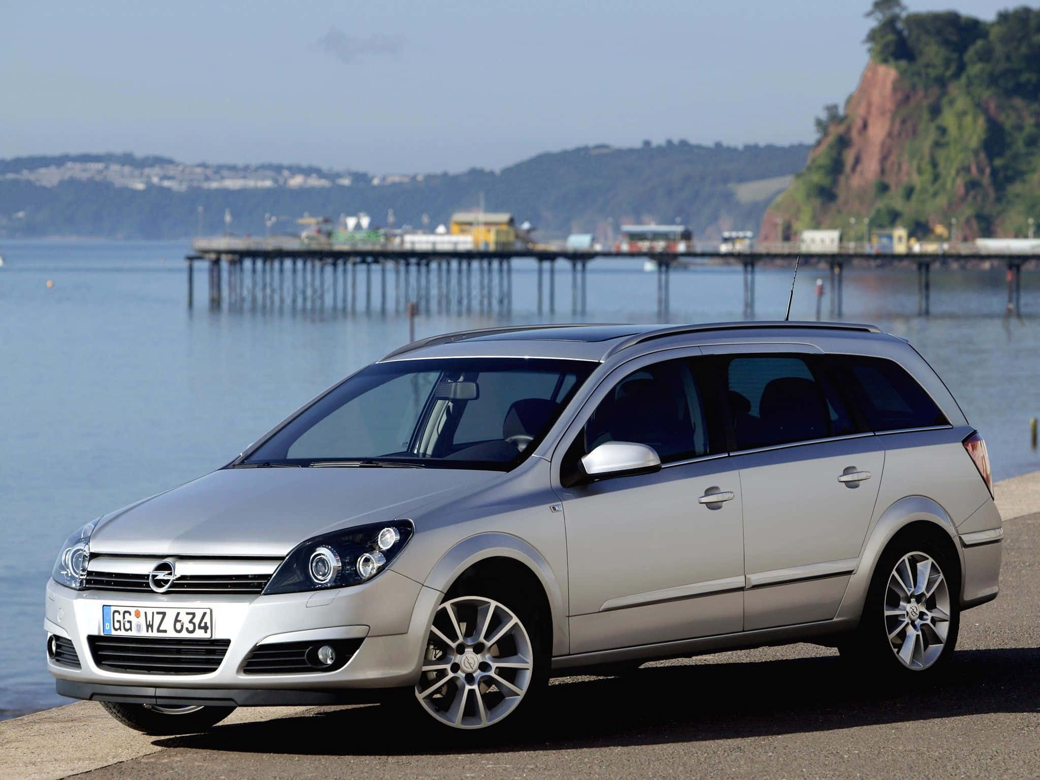 Opel Astra 桌布 壁紙 — 一輛銀色旅行車停泊在海濱，背景有碼頭和懸崖，天空晴朗。
