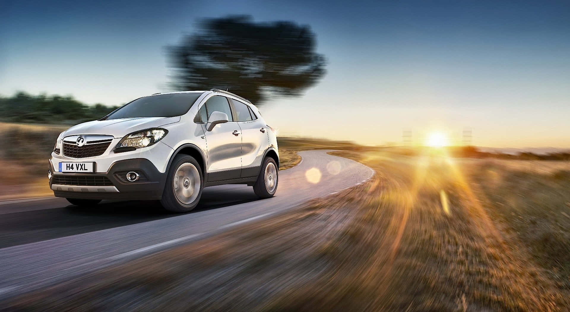 Opel Mokka 桌布 壁紙 — 一輛銀色SUV在日落時分沿著蜿蜒的道路疾馳，伴隨著動態模糊和太陽散發出的金色光芒。