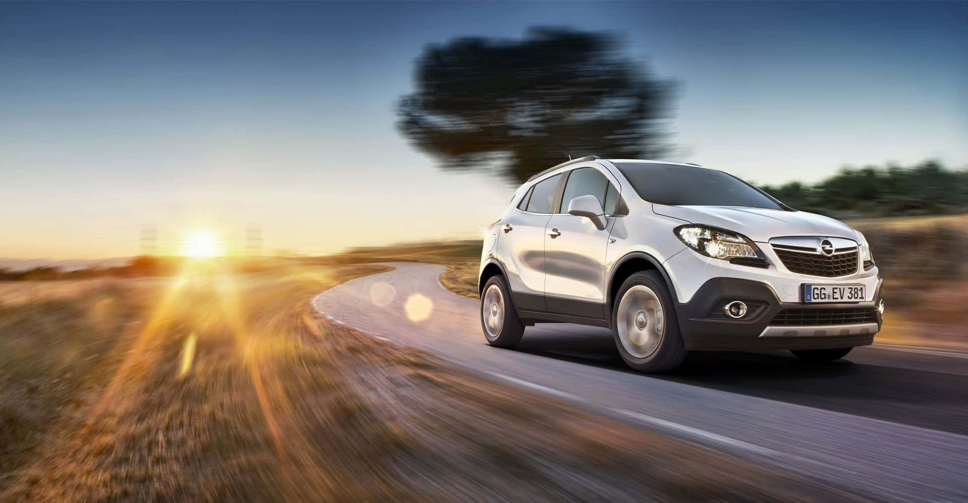 Opel Mokka 桌布 壁紙 — 一輛銀色SUV在日落時分沿著蜿蜒的道路疾馳，帶有動態模糊和太陽散發的金色光芒。