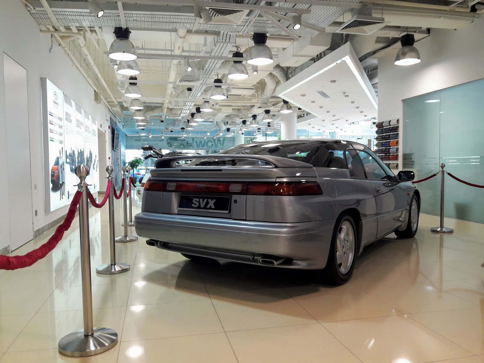 Subaru Svx 桌布 壁紙 — 一輛銀色SVX跑車在現代化展廳中展示，配有紅色繩索屏障和明亮的頂燈照明。