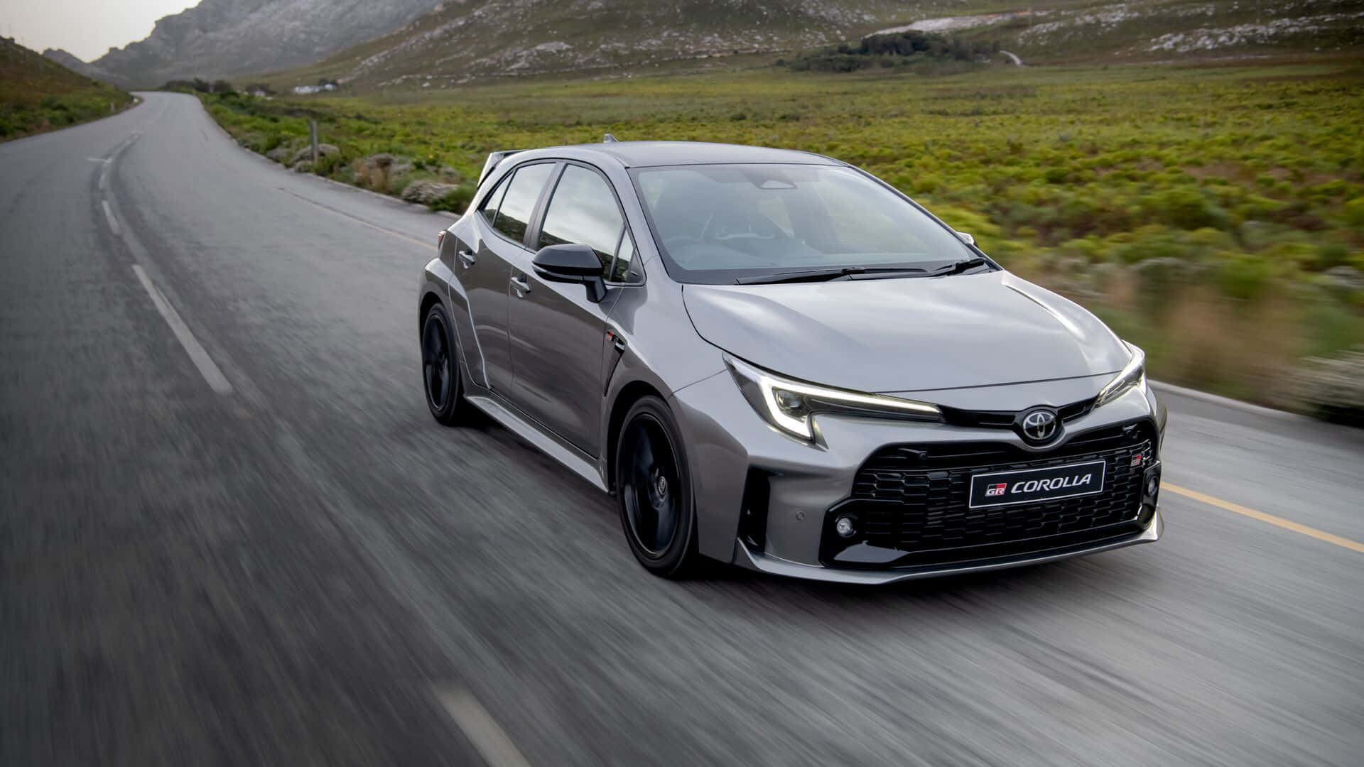  壁紙 — 一輛流線型的銀色豐田 Corolla GR Sport 在蜿蜒的山路上穿過綠色山丘飛馳。