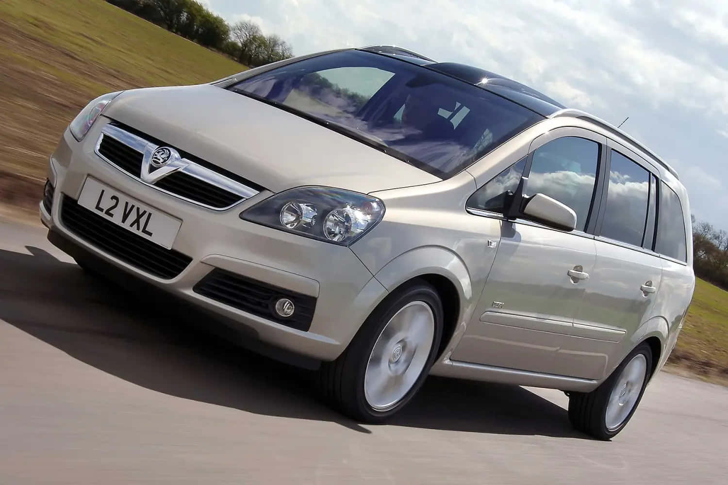 Opel Zafira 桌布 壁紙 — 一輛銀色嘅Vauxhall Zafira喺一條有模糊草地同多雲天空做背景嘅路上行駛。