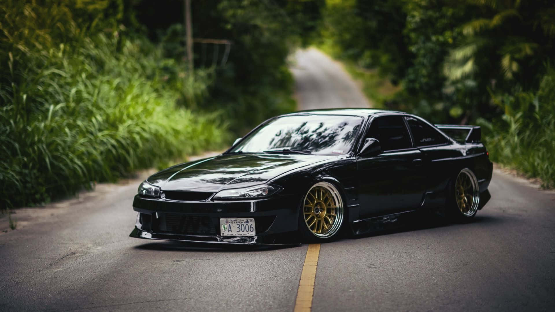 日產Silvia S15金色鋁圈 壁紙 — 一輛時尚的黑色跑車配金色輪圈，停在蜿蜒的森林道路上，周圍綠意盎然。