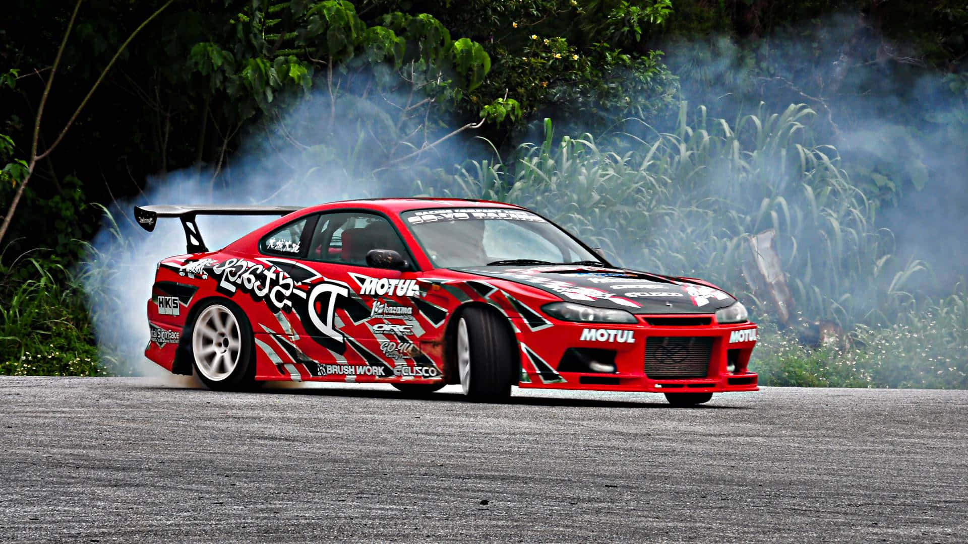 夜空下流暢迅捷的尼桑Silvia S15 壁紙 — 一輛帶有黑色圖案的紅色飄移車在鋪砌的道路上側滑，周圍是茂密的綠色樹葉，冒出濃煙。