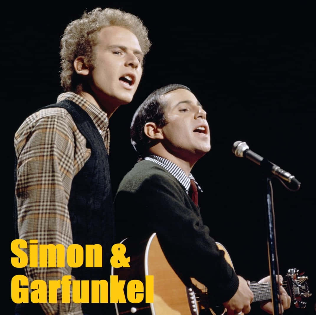  壁紙 — Simon & Garfunkel 在舞台上表演，Garfunkel 對著咪高峰唱歌，Simon 在戲劇性的舞台燈光下彈結他。