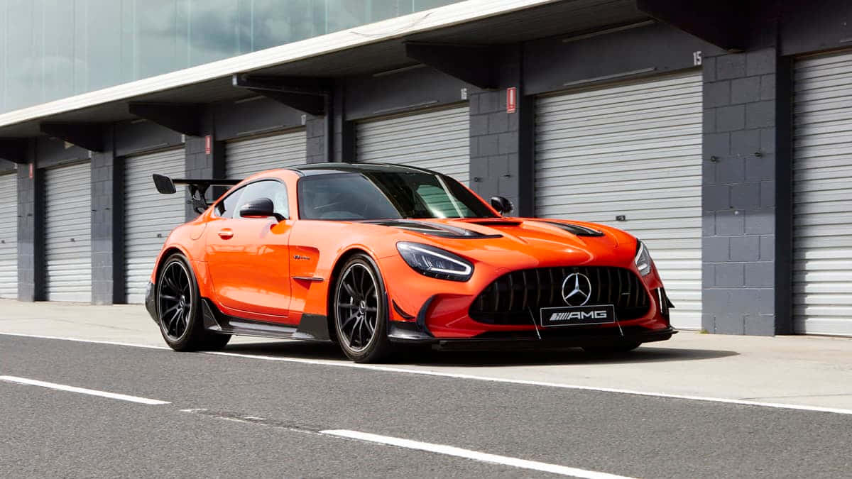 Mercedes Benz Amg Gt 桌布 壁紙 — 一輛鮮橙色的 Mercedes-AMG GT R 跑車在明亮的日光下停放在工業車庫門前。