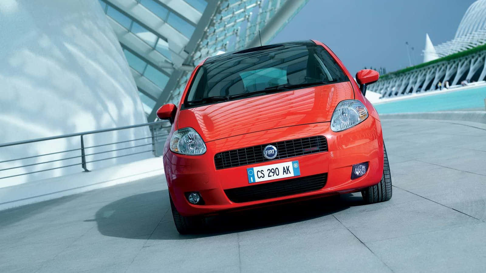 Fiat Punto 桌布 壁紙 — 一輛鮮紅色的Fiat Punto停泊在現代混凝土廣場上，背景是晴朗的藍天和時尚的建築結構。