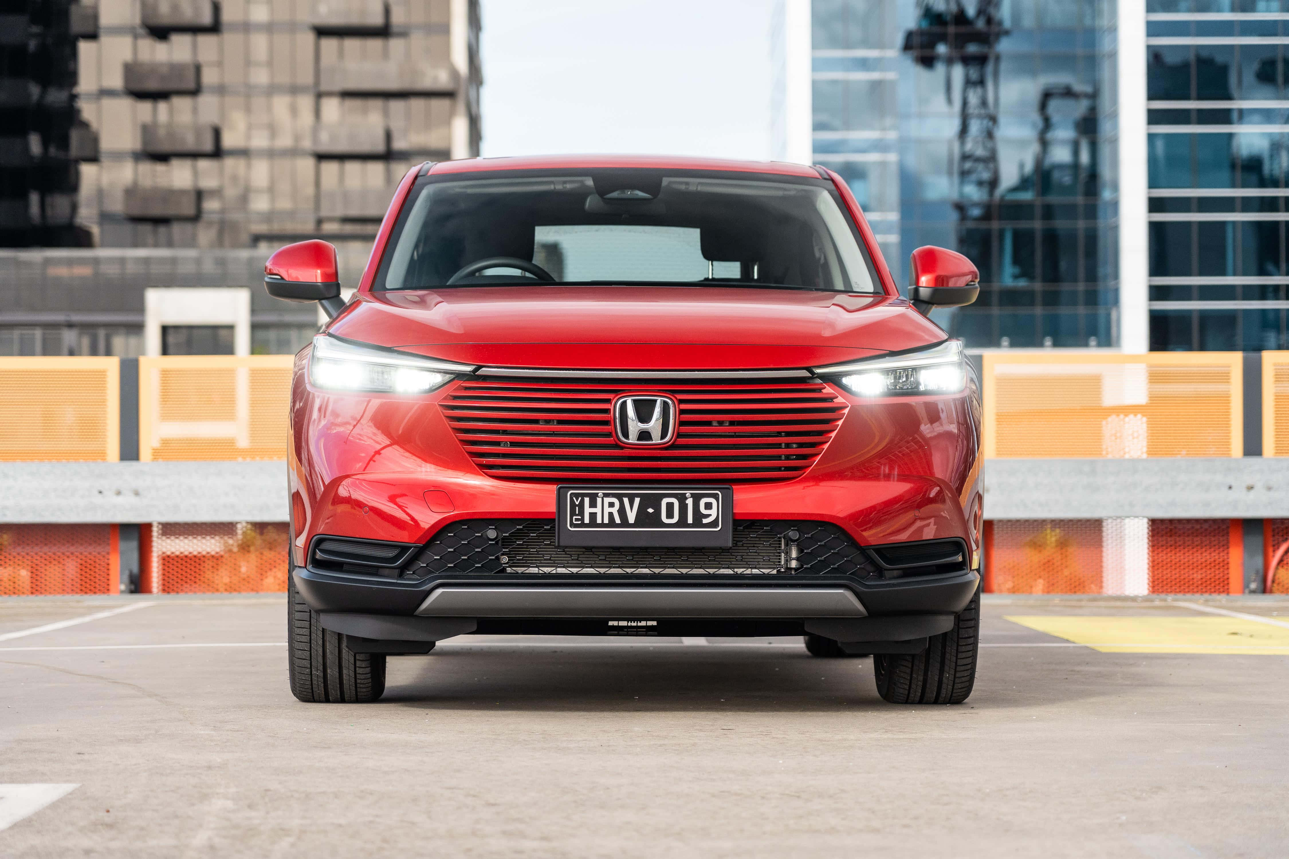Honda Hr-v 桌布 壁紙 — 一輛鮮紅色的本田HRV電動SUV在現代城市停車場中向前，背後有玻璃摩天大樓。