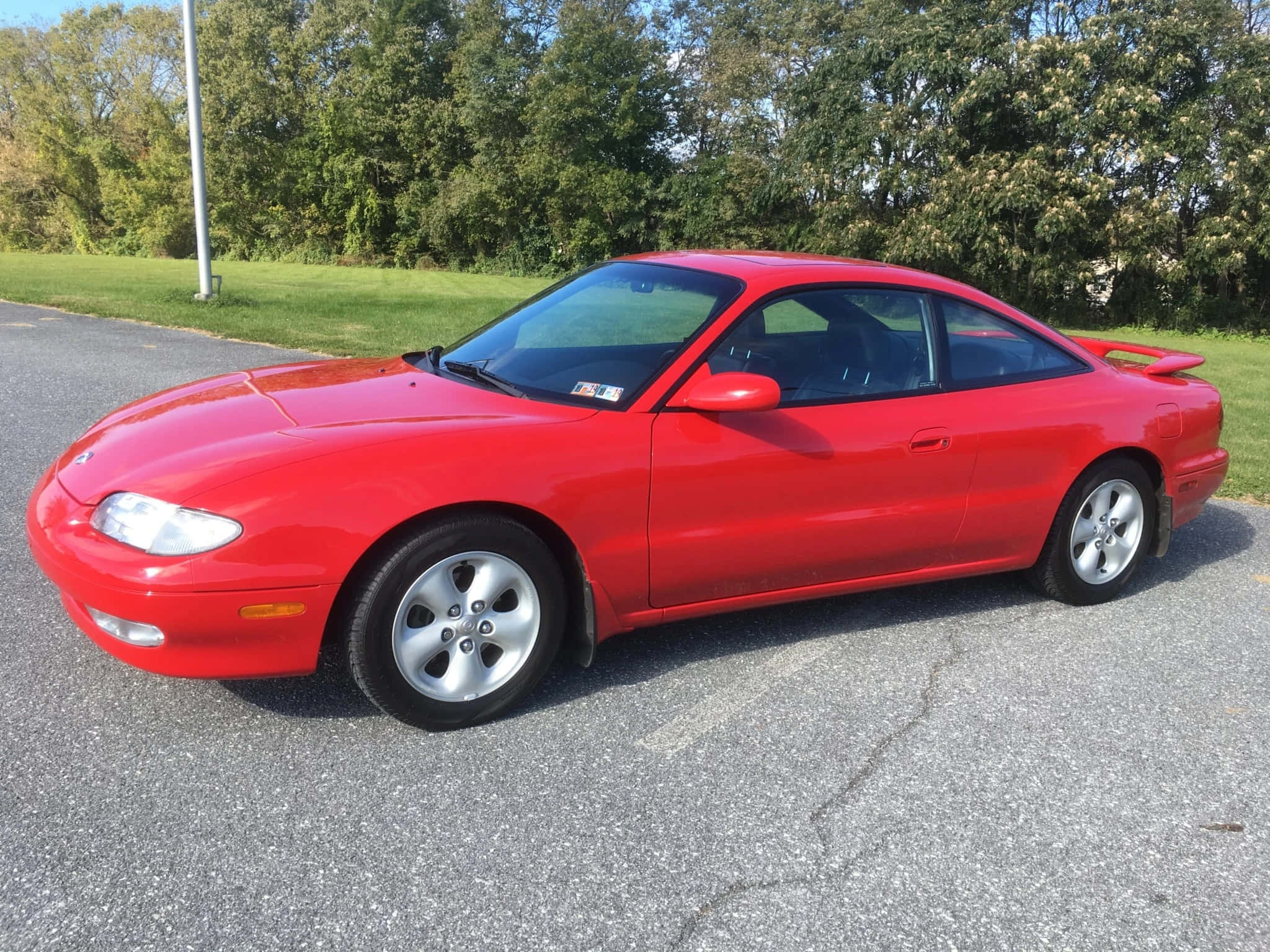 Mazda Mx-6 桌布 壁紙 — 一輛鮮紅色的萬事得MX-5跑車停在裂開的瀝青路上，背景有綠樹和草地，在明亮的日光下。