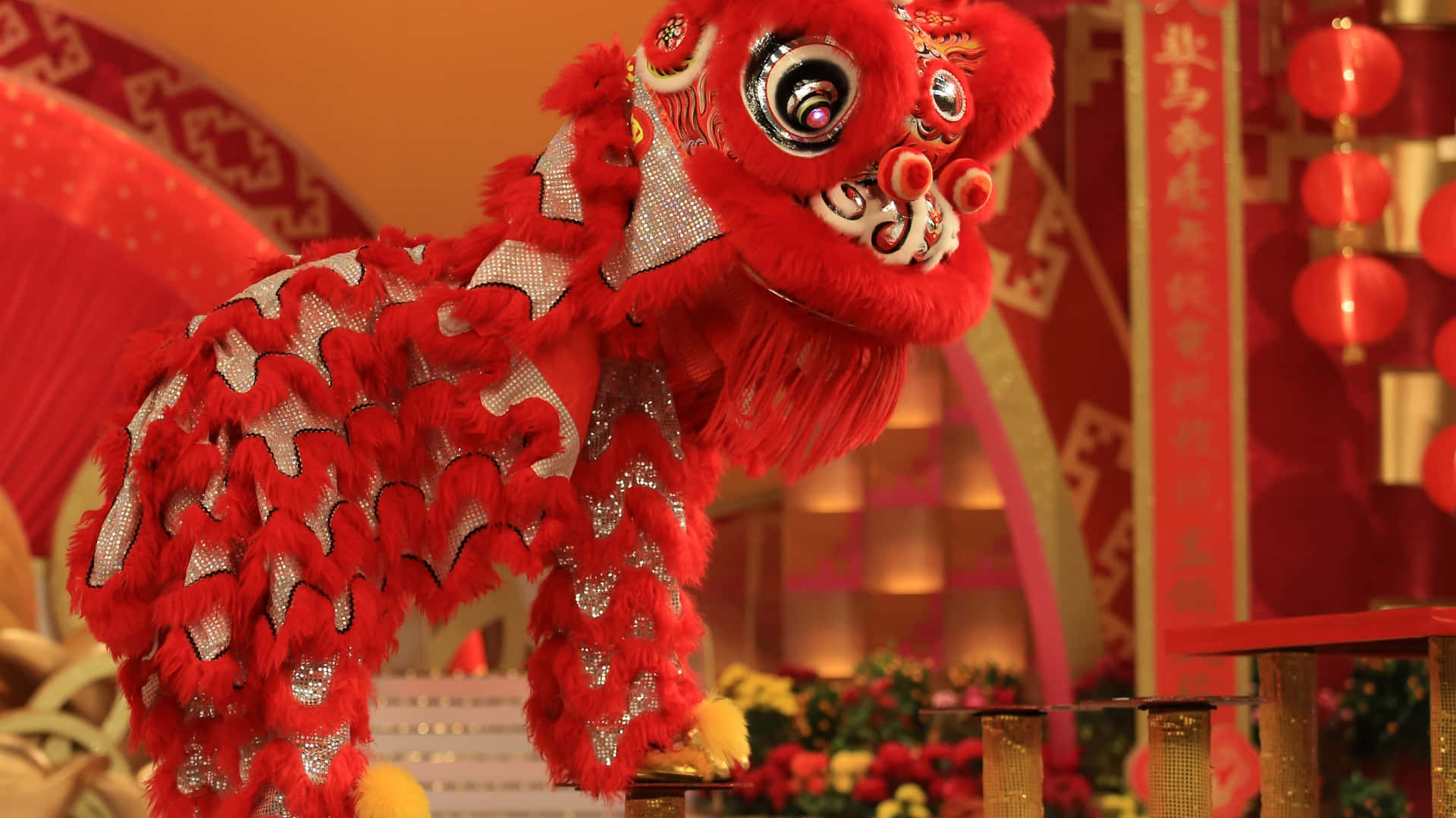  壁紙 — 一件鮮紅色嘅舞獅服裝，帶有閃爍嘅銀色點綴，喺掛滿燈籠同傳統裝飾嘅節日背景下表演。