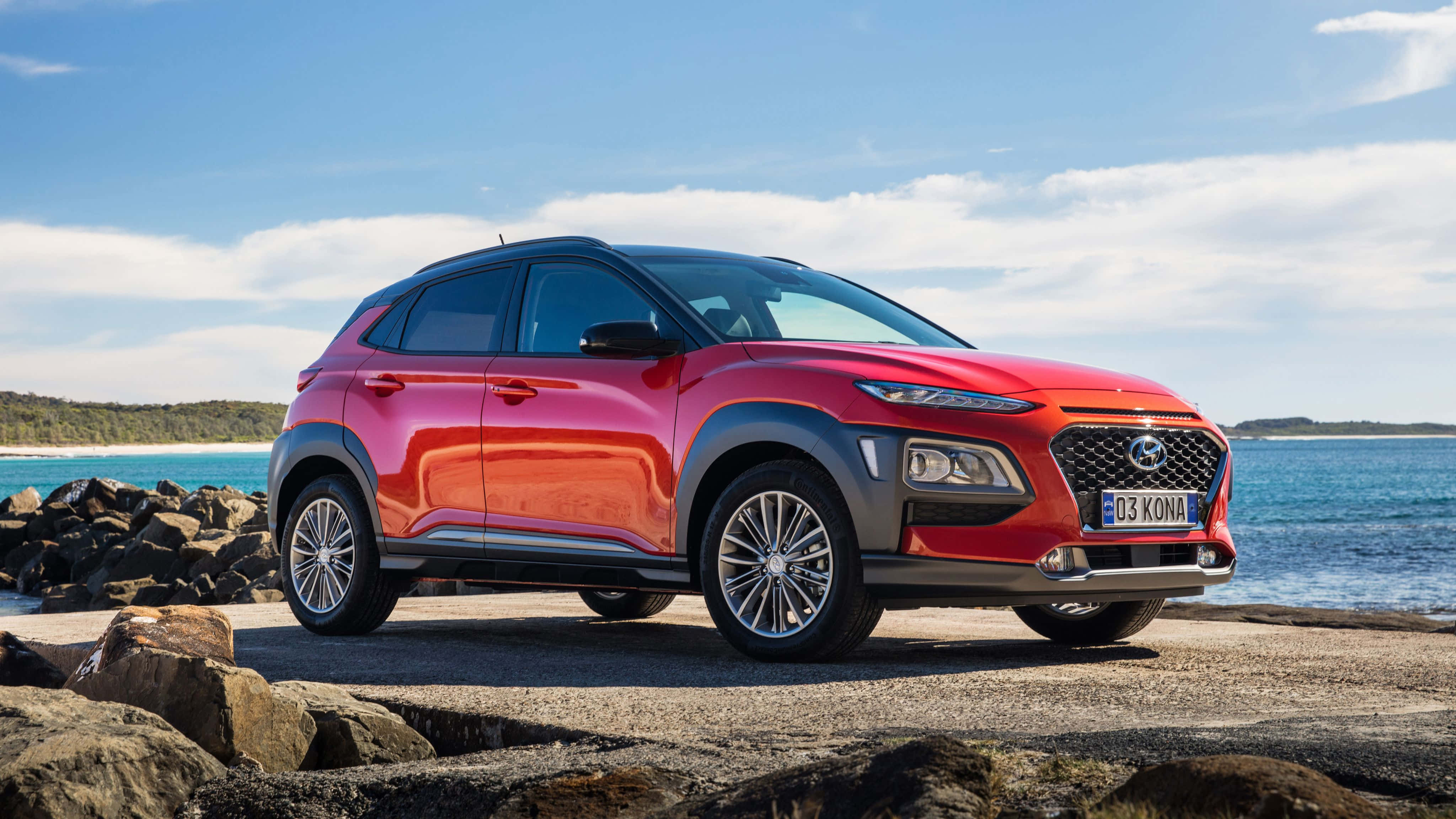 Hyundai Kona 桌布 壁紙 — 一輛鮮紅色的現代Kona SUV停泊在岩石海岸道路上，背後是碧綠色的海洋和清澈的藍天。