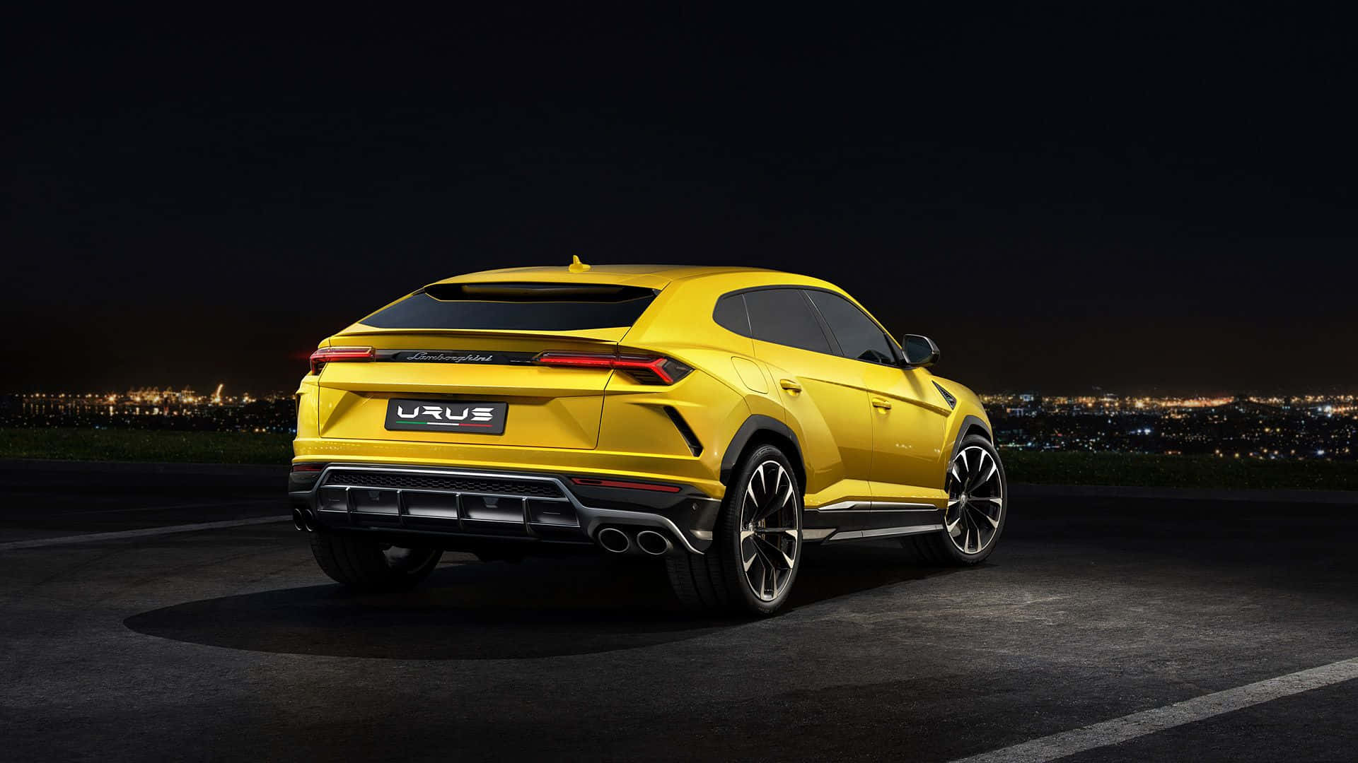 Lamborghini Urus 桌布 壁紙 — 一輛鮮黃色的林寶堅尼 Urus SUV 在夜間停泊，遠處城市燈光閃爍。