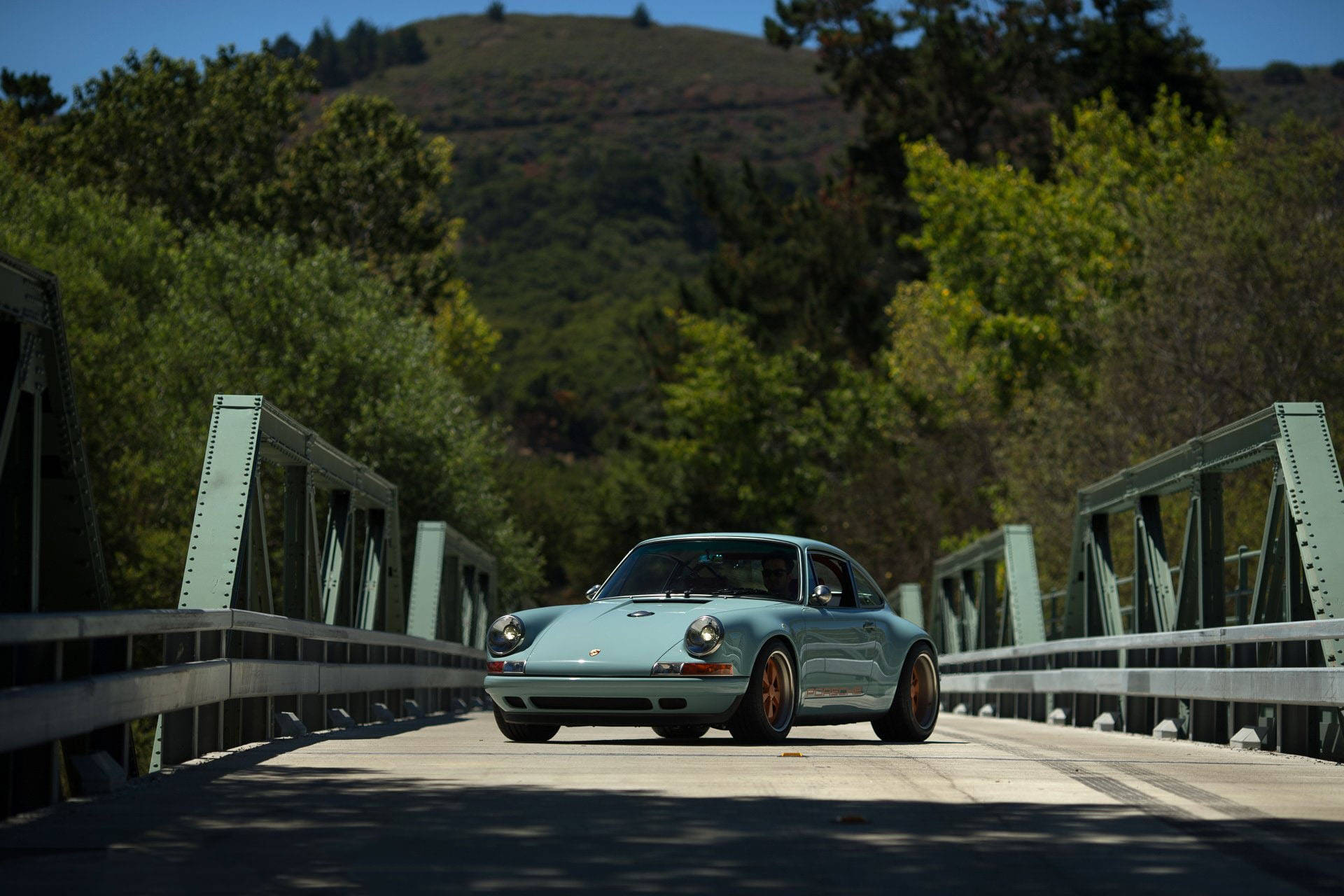 Singer Porsche靜候於古橋之上 壁紙 — 一輛淺藍色經典保時捷911在晴朗藍天下駛過綠色金屬橋，背景是森林覆蓋的山丘。