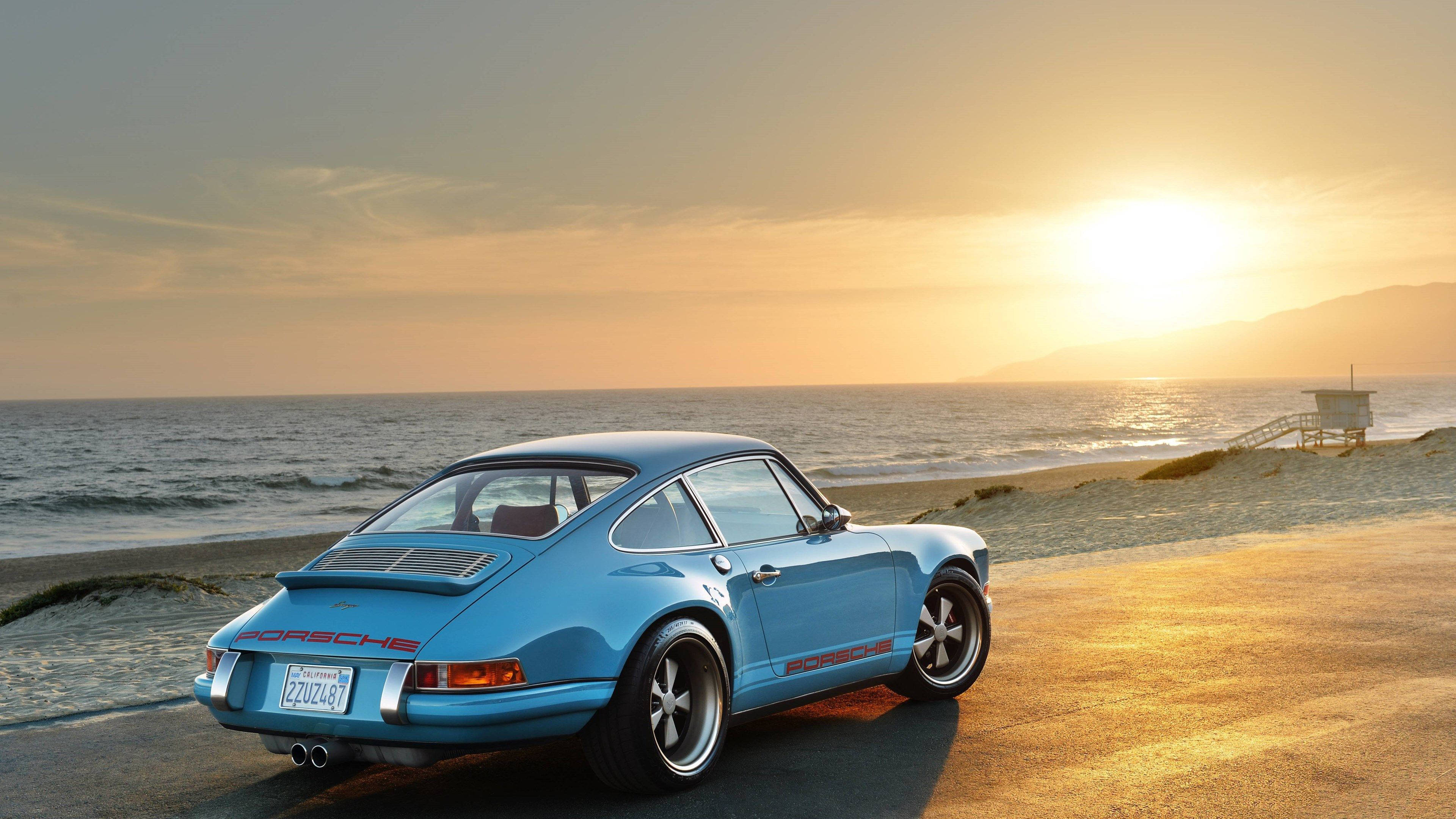 Singer Porsche於海灘日落 壁紙 — 一架淺藍色經典保時捷911停泊在日落時分的海岸公路上，背景有海浪和救生塔。