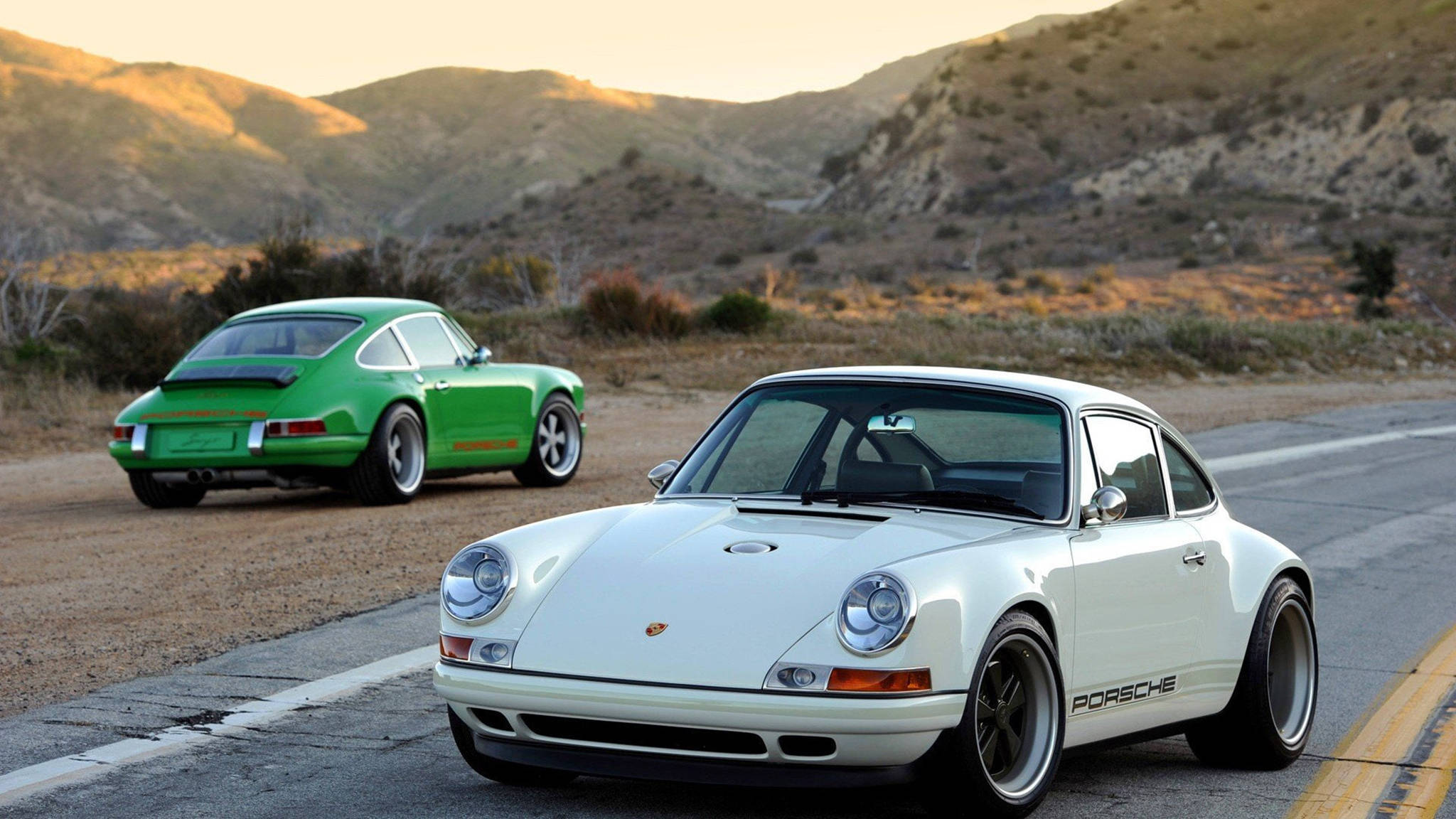 Singer Porsche車隊 壁紙 — 兩輛經典保時捷911，一白一綠，停泊在沙漠公路上，起伏的山丘沐浴在金色的日落光芒中。