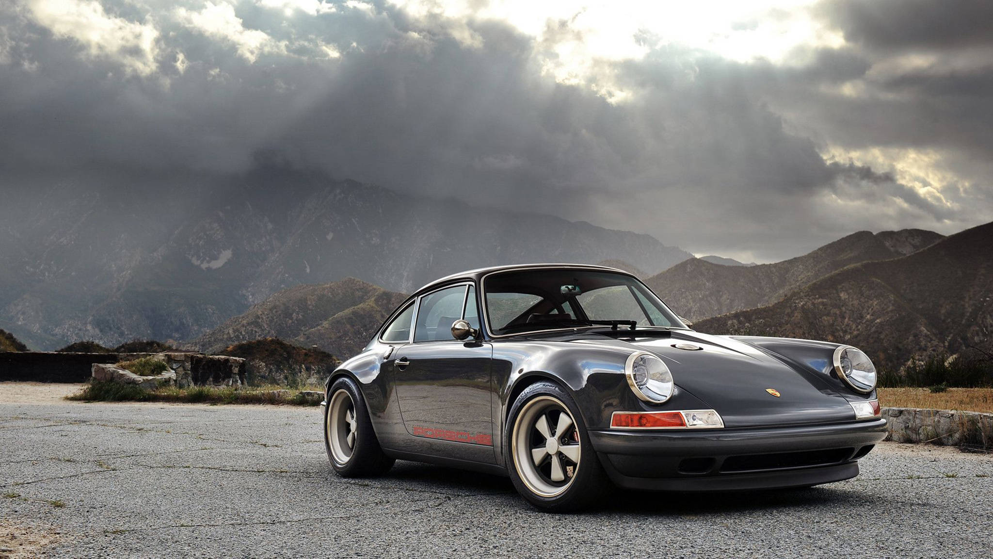 Singer Porsche陰天背景 壁紙 — 一架時尚黑色保時捷911跑車停泊在碎石路上，背景是風暴天空下的戲劇性山脈。