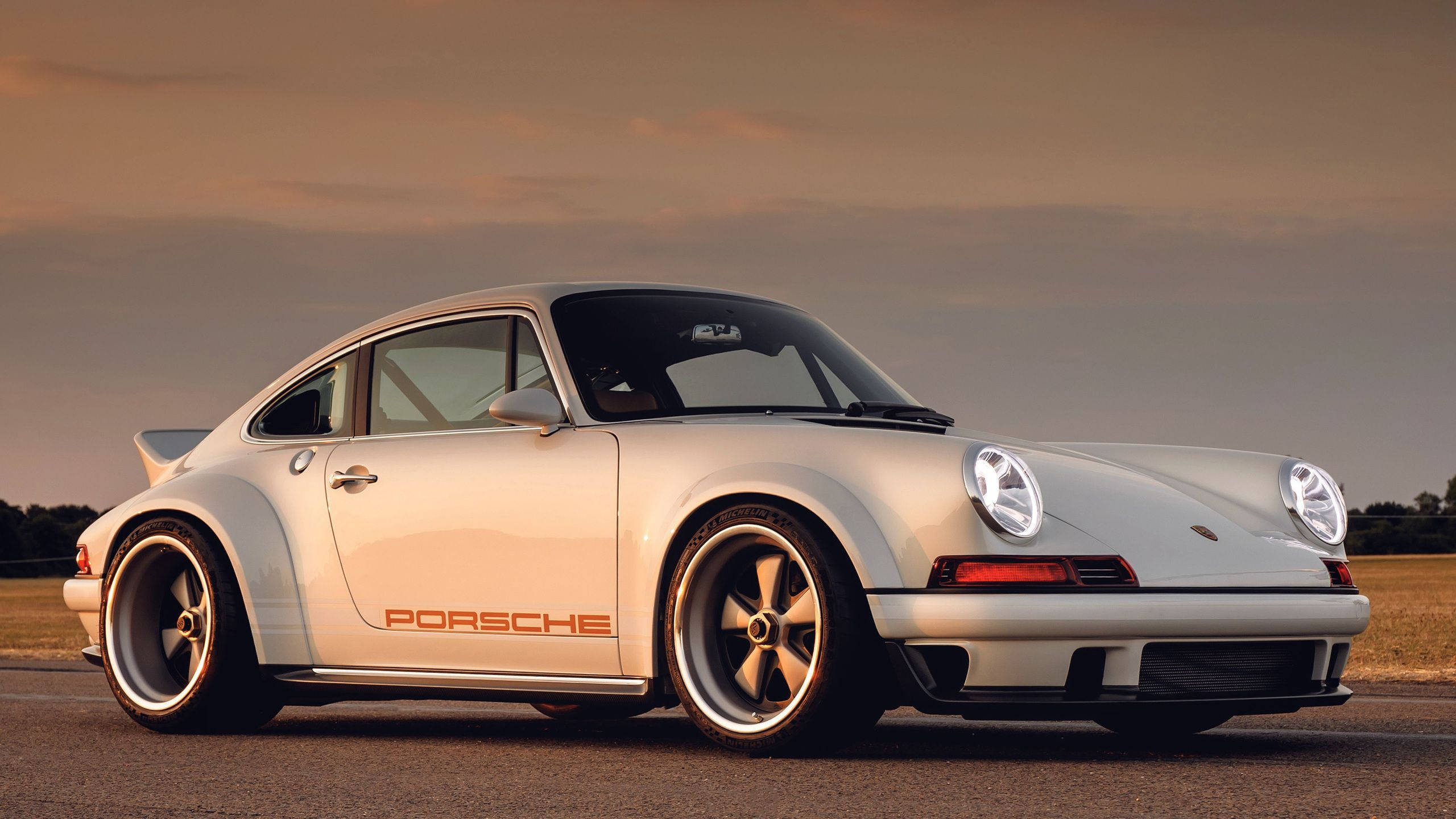 Singer保時捷911的優雅美感 壁紙 — 一架時尚的白色保時捷911經典跑車停泊在瀝青路上，正值黃金時段，溫暖的夕陽照明。