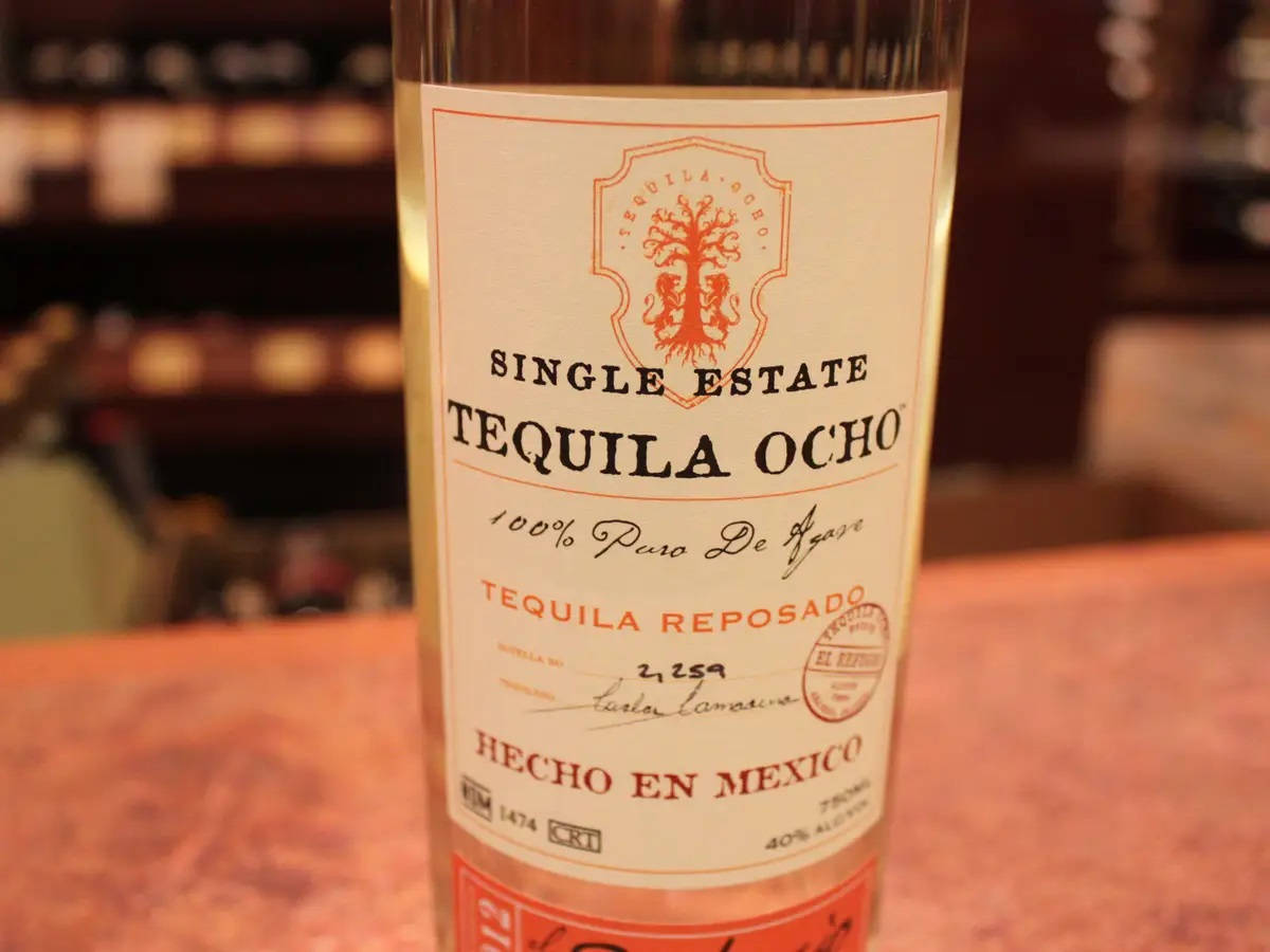Single Estate Tequila Ocho 瓶身標籤特寫 壁紙 — 單一莊園龍舌蘭酒瓶的特寫，標籤為「Tequila Ocho」，放在木吧檯上，背景有模糊的架子。