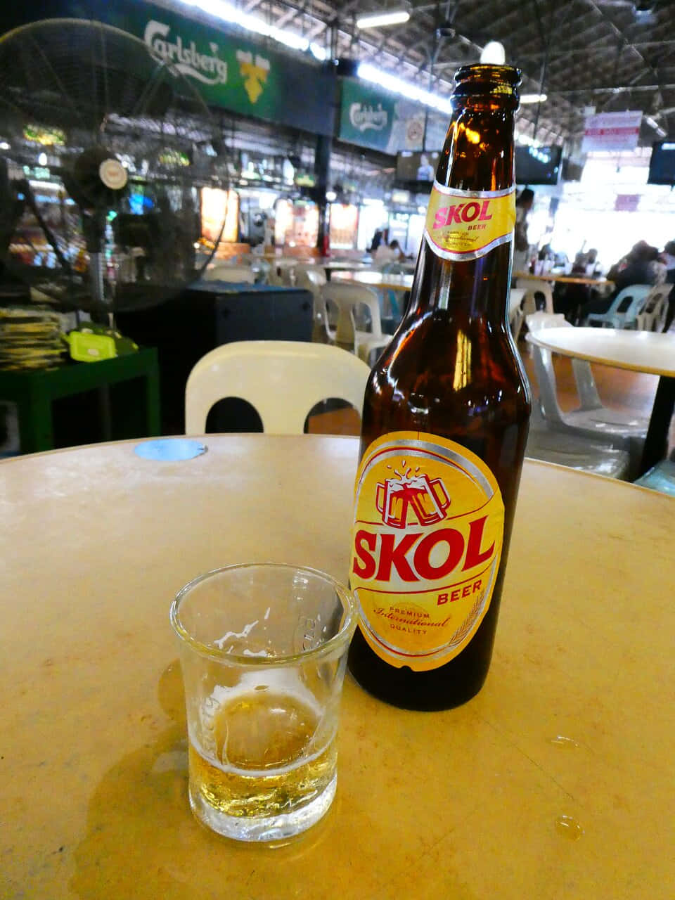  壁紙 — 一個棕色Skol啤酒瓶和半滿的玻璃杯放在黃色桌子上，室內食肆內有風扇和顧客，氣氛熱鬧。