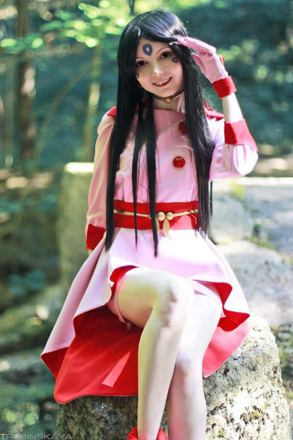 Skuld 女神 cosplay 自然風景 壁紙 — 一位穿著粉紅色和紅色服裝、額頭上有封印的角色扮演者，微笑著坐在陽光普照的森林中長滿苔蘚的石頭上。