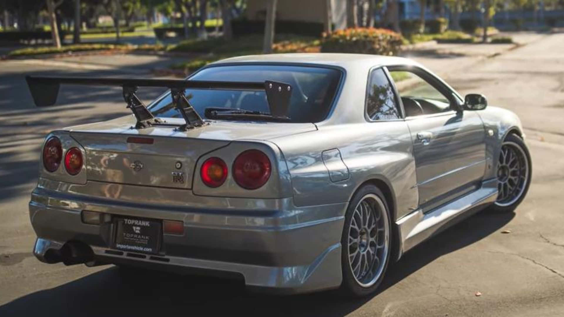 保羅·沃克駕駛日產Skyline撕裂柏油路面 壁紙 — 一架銀色日產Skyline R34跑車，帶有大型尾翼，停泊在陽光普照的郊區街道上。