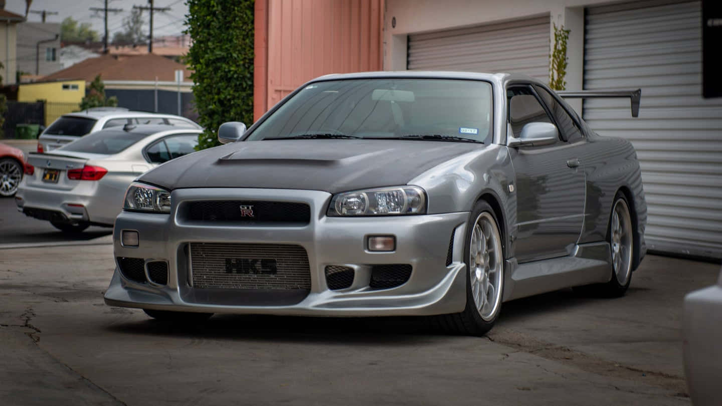 保羅·沃克駕駛他的Skyline跑車 壁紙 — 一輛帶有HKS標誌的時尚銀色日產Skyline R34停泊在車庫旁的混凝土車道上。