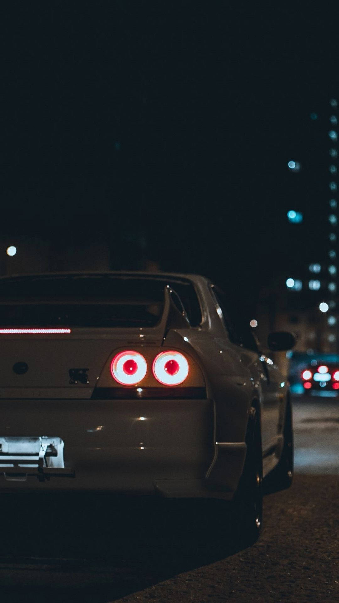 日產Skyline GT-R iPhone汽車 壁紙 — 一輛銀色跑車，帶有發光的紅色圓形尾燈，停泊在夜晚黑暗的城市街道上，背景有模糊的燈光。