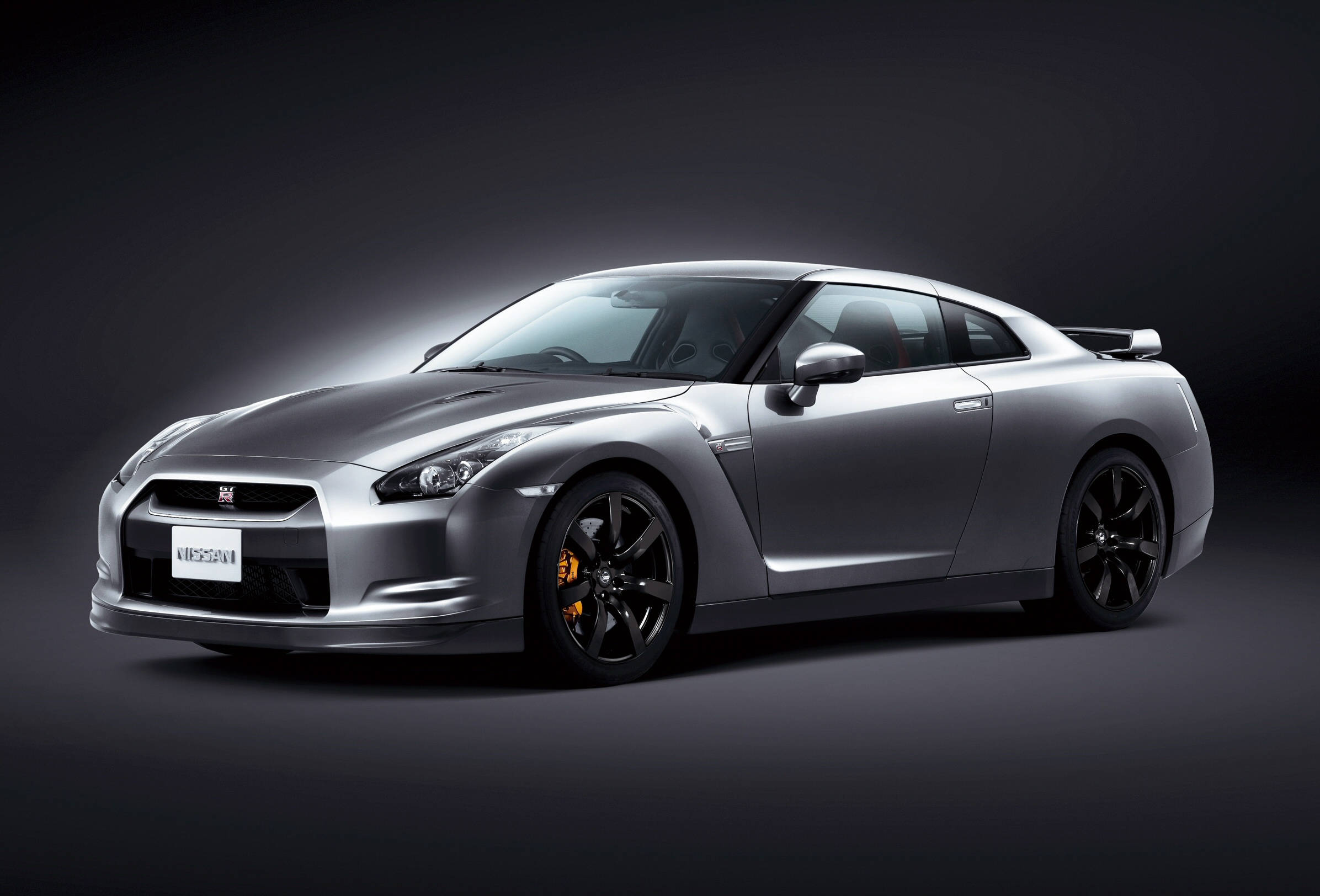尼桑Skyline GTR R35深灰色款 壁紙 — 一輛時尚的銀色日產GT-R跑車，帶有黑色飾邊和橙色制動卡鉗，在深色工作室背景下擺拍。