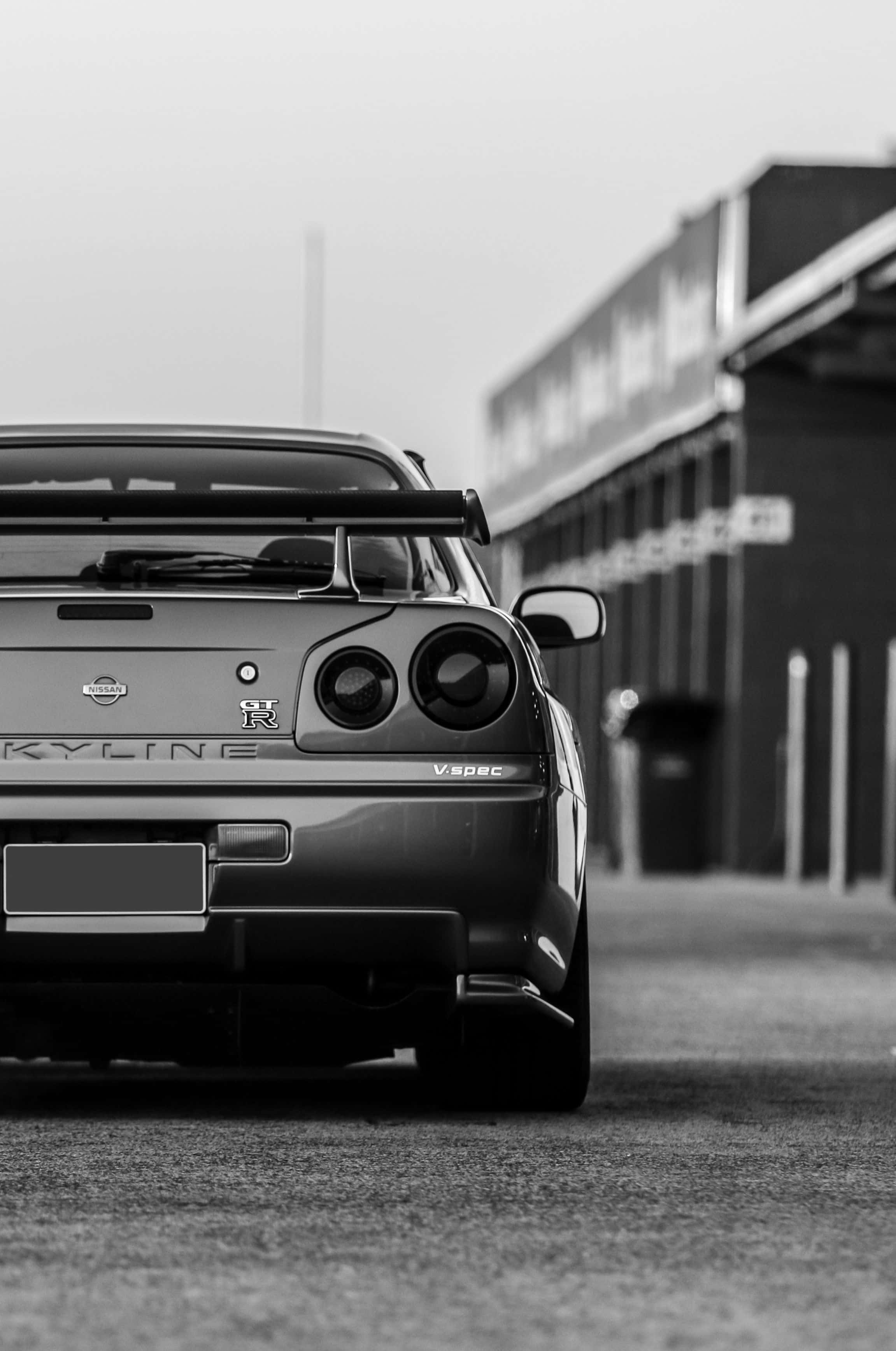 黑白風格尼桑Skyline iPhone壁紙 壁紙 — 一輛日產Skyline GT-R V-SPEC停泊在車庫建築物前的黑白背面視圖。
