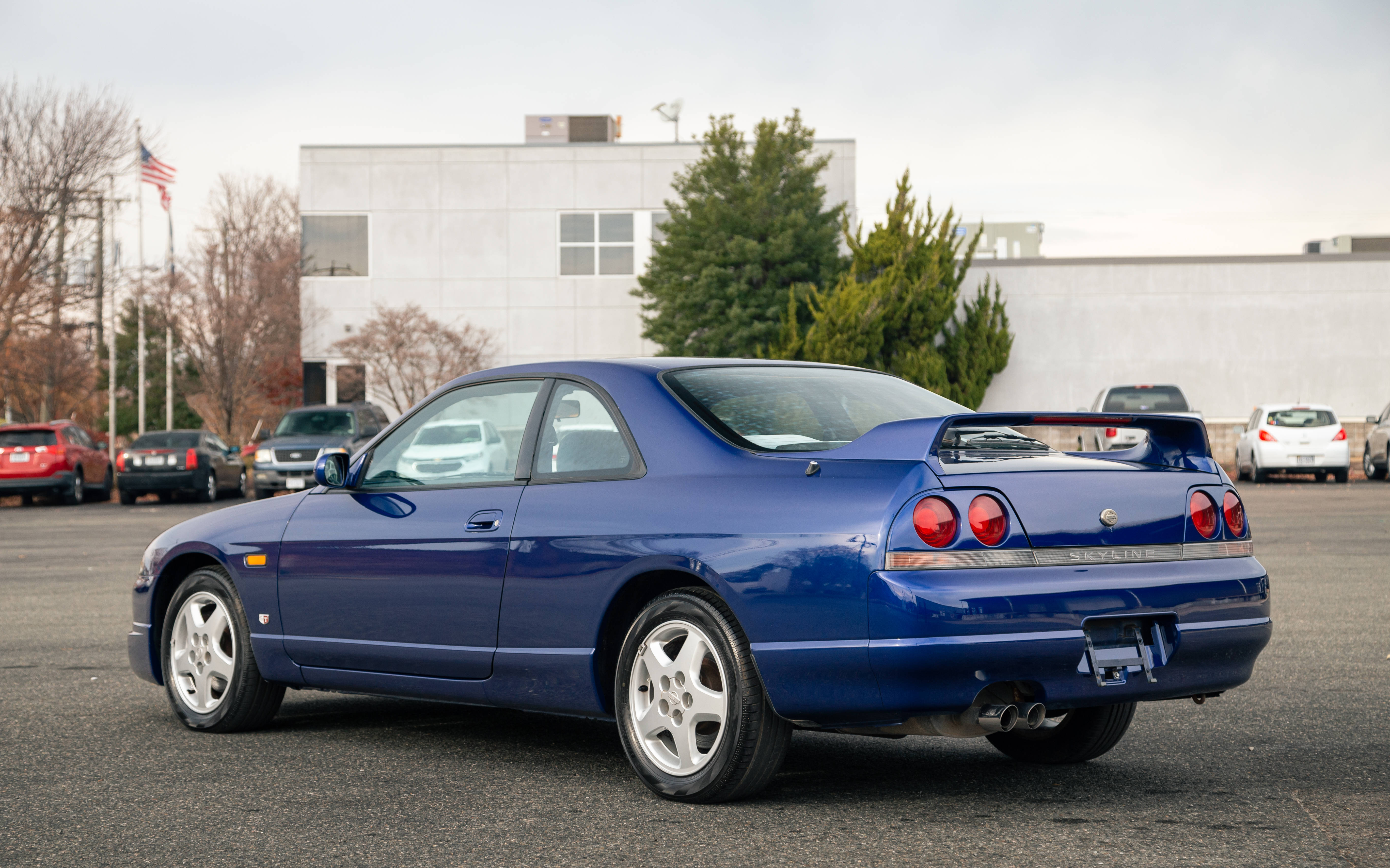藍天下的無牌Skyline賽車 壁紙 — 一輛充滿活力的藍色日產Skyline GT-R雙門跑車停泊在陰天的地方，背景有光禿禿的樹木和現代建築。