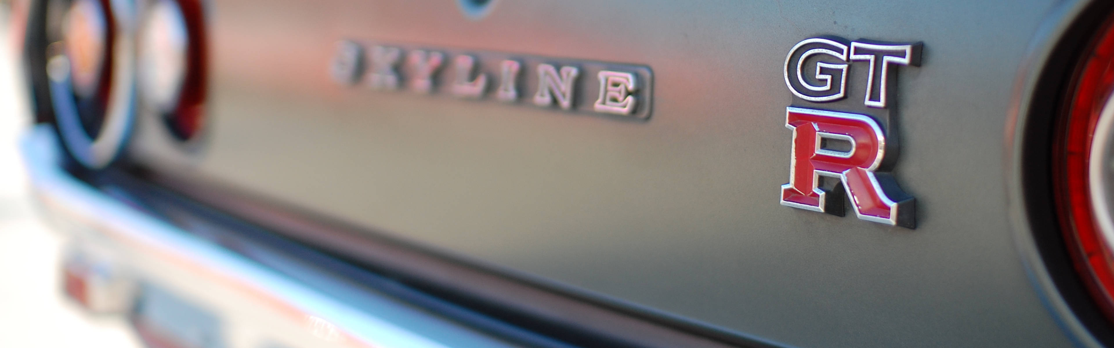 Skyline 車尾標誌特寫 壁紙 — 金屬灰色日產Skyline GT-R徽章的近距離特寫，帶有紅色和銀色字樣，兩側是圓形尾燈。