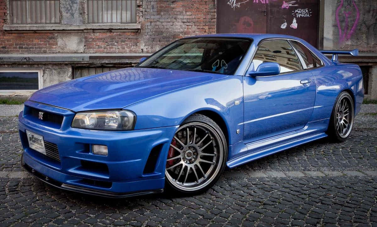 保羅·沃克駕駛日產Skyline參與街頭賽車 壁紙 — 一輛充滿活力的藍色日產Skyline R34跑車，配備定制輪圈，停在鵝卵石街道上，旁邊是一棟佈滿塗鴉的磚砌建築。