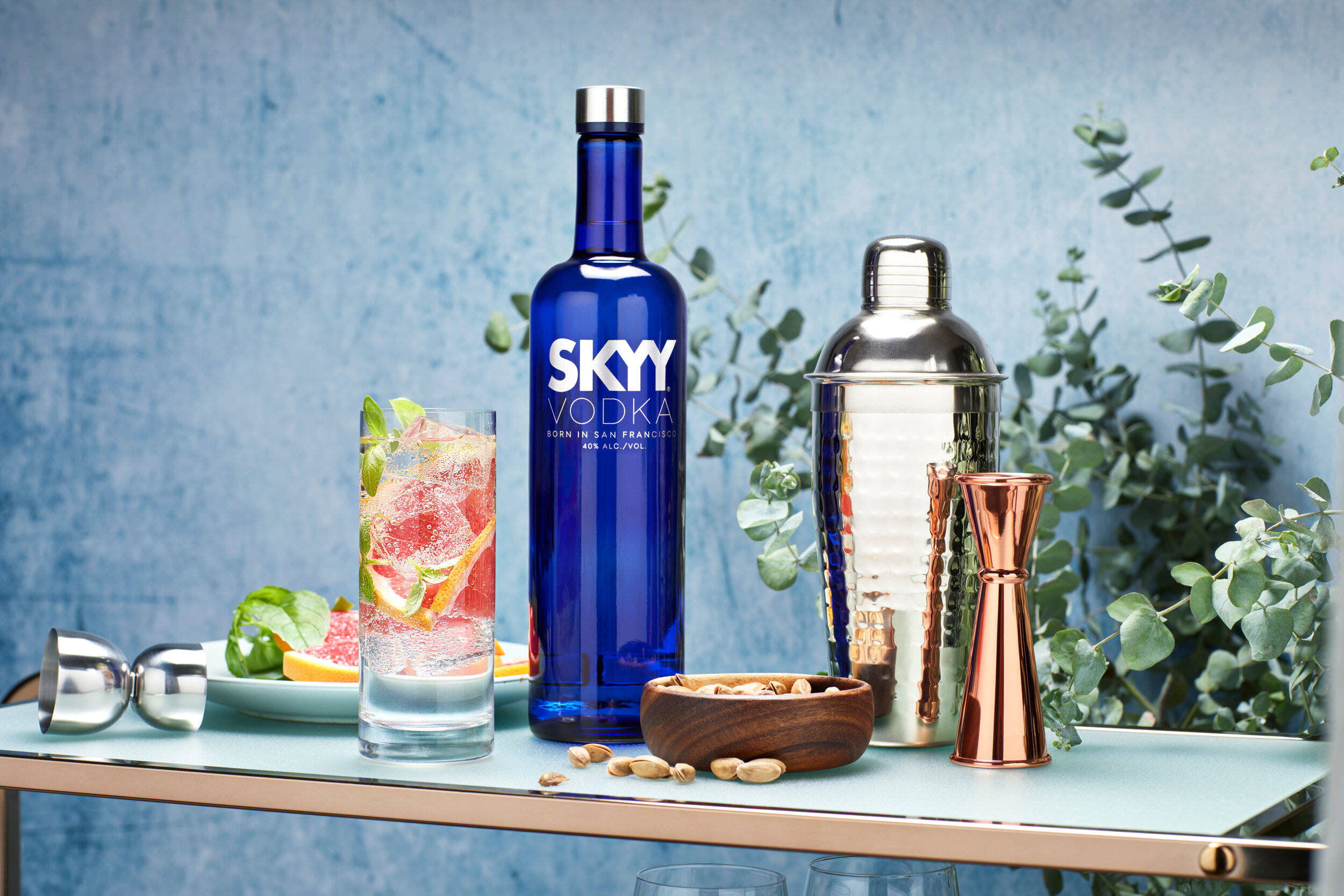 Skyy伏特加配雞尾酒 壁紙 — 一個充滿活力的藍色Skyy伏特加酒瓶旁邊放著一個裝飾有柑橘和香草的雞尾酒杯，放在一個時尚的吧檯上，配有銅和銀的吧檯工具以及桉樹葉。
