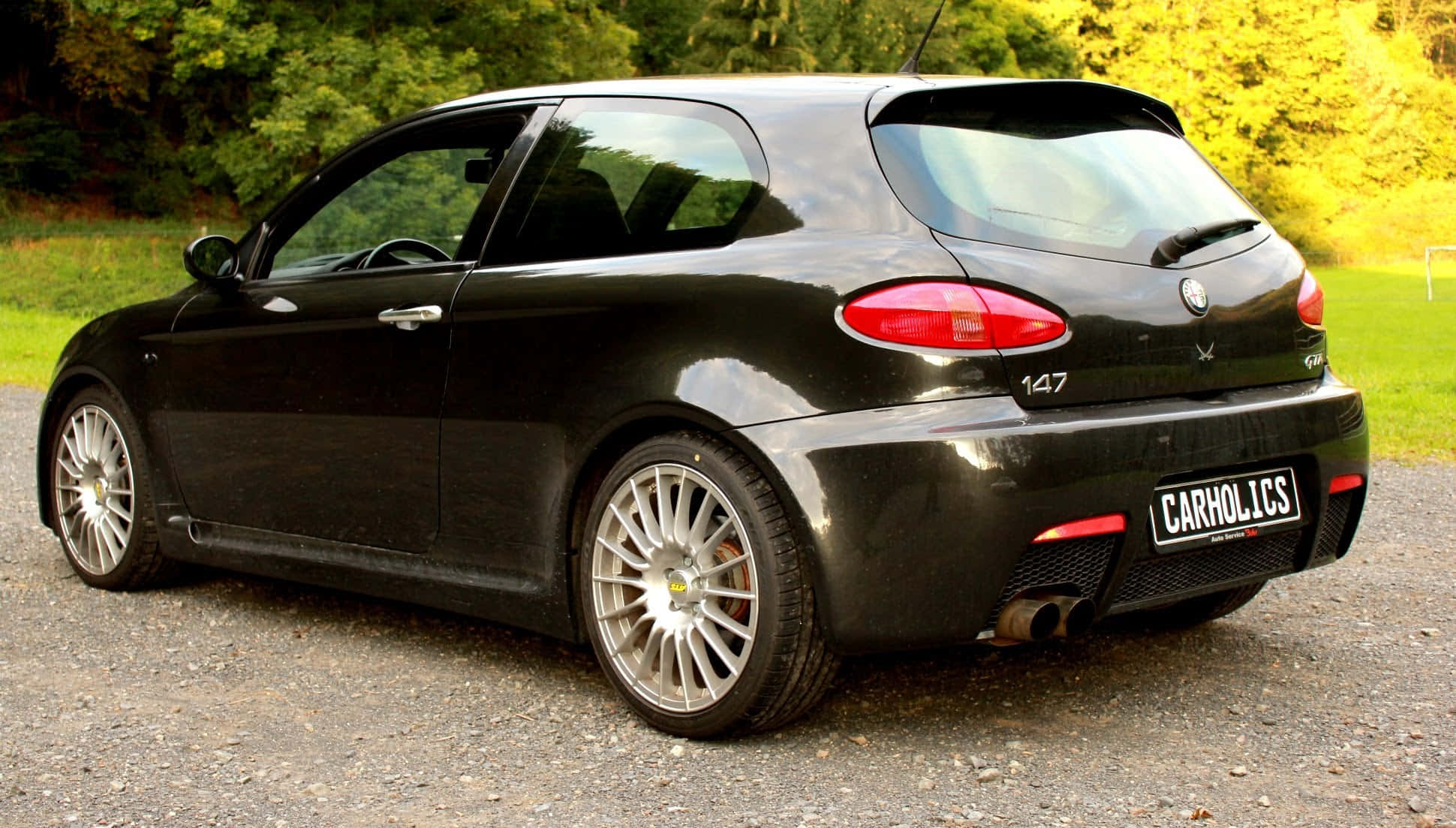 Alfa Romeo 147 桌布 壁紙 — 一輛時尚的黑色愛快羅密歐147跑車停在碎石上，背景是茂密的綠樹和草地。