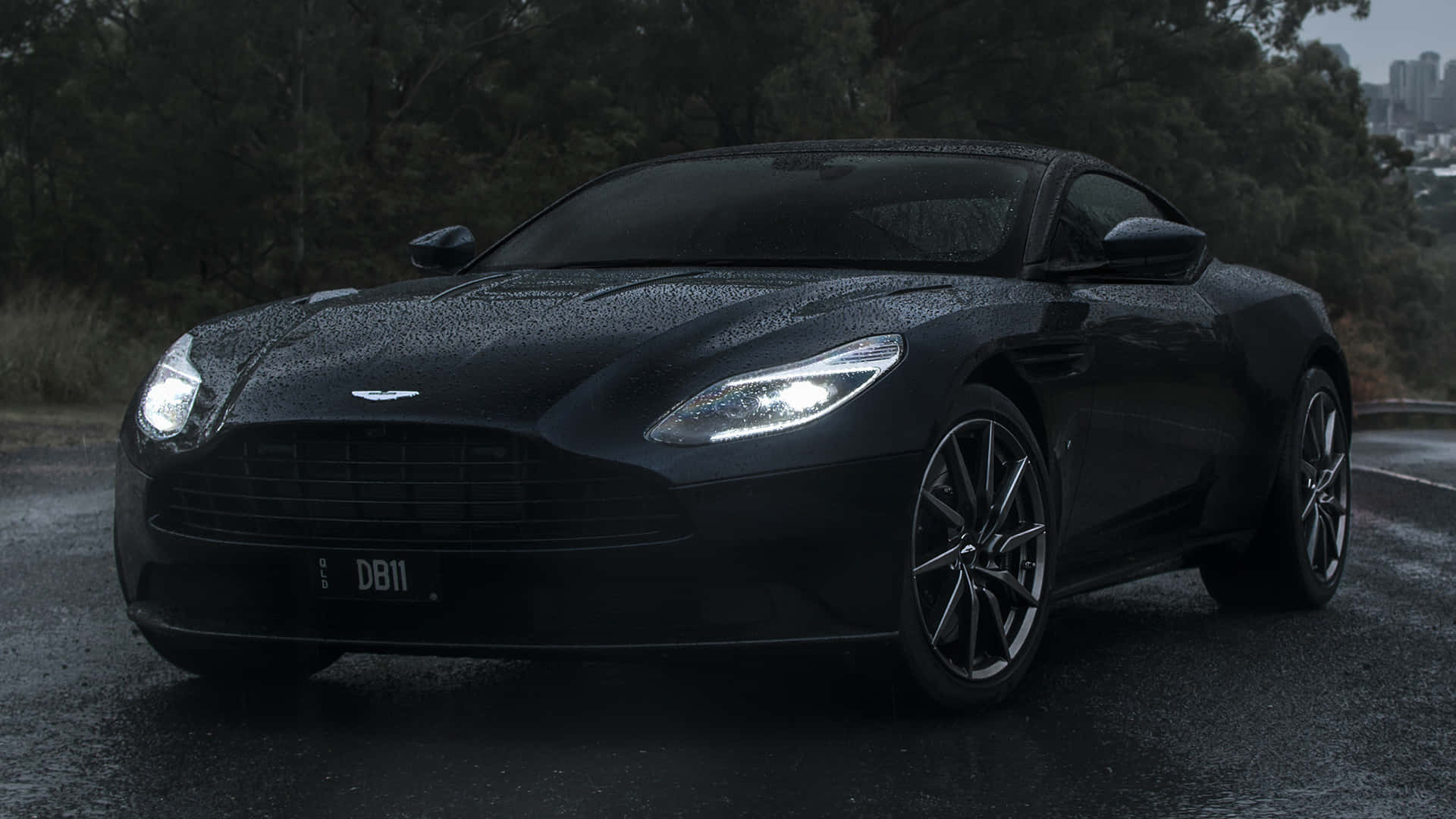 Aston Martin Db11 桌布 壁紙 — 一輛時尚的黑色Aston Martin DB11在雨中閃耀，車頭燈亮著，停在濕漉漉的道路上，旁邊有樹木和遠處的城市天際線。