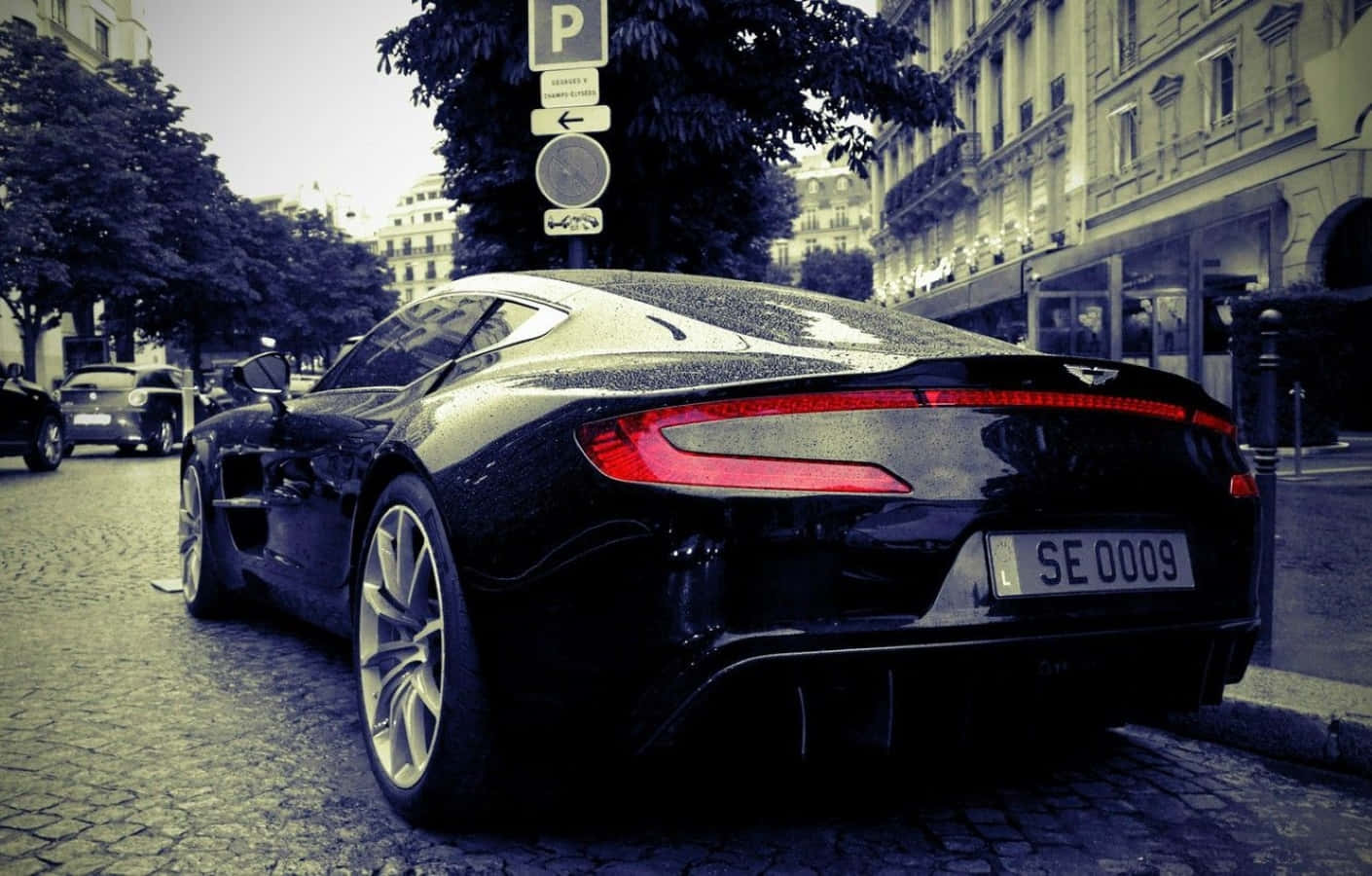 Aston Martin One-77 桌布 壁紙 — 一架型格黑色Aston Martin跑車停泊在濕漉漉的鵝卵石街道上，背景是復古歐洲建築。