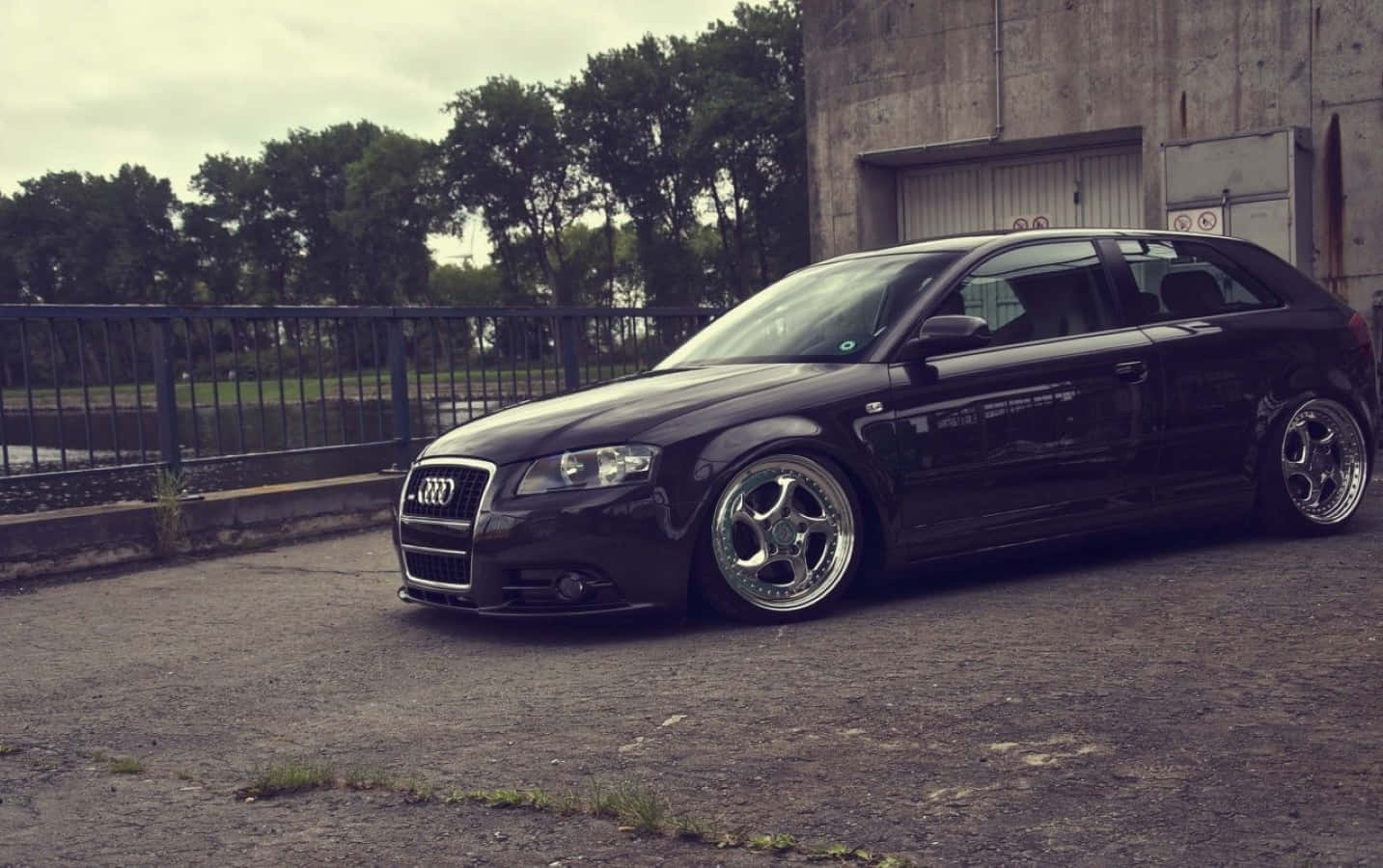 Audi A3 桌布 壁紙 — 一輛時尚的黑色奧迪旅行車，配備定制鍍鉻輪圈，停在裂縫的瀝青路上，旁邊是生鏽的圍欄和混凝土建築，天空多雲。