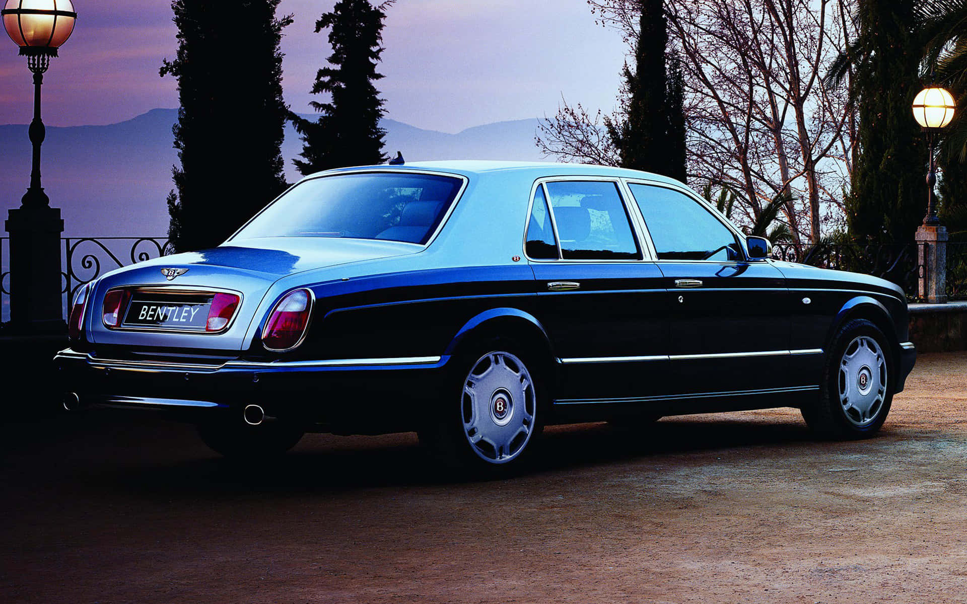 Bentley Arnage 桌布 壁紙 — 一輛時尚的黑色賓利轎車在暮色燈光下閃耀，背景是高聳的樹木和遠山。
