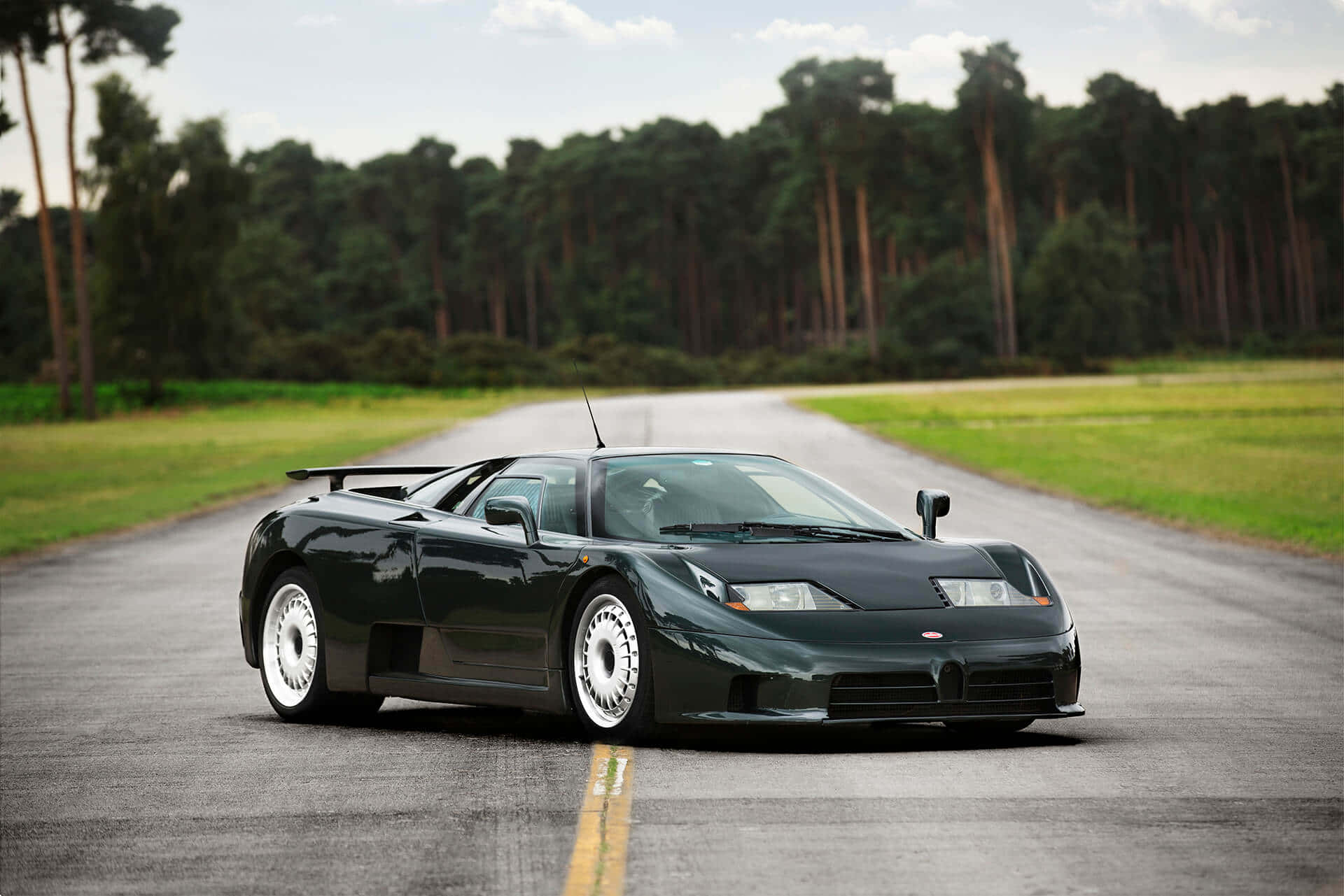 Bugatti Eb110 桌布 壁紙 — 一部時尚的黑色布加迪EB110超級跑車在蜿蜒的瀝青路上行駛，兩旁是茂密的綠色田野和茂密的森林，天空部分多雲。