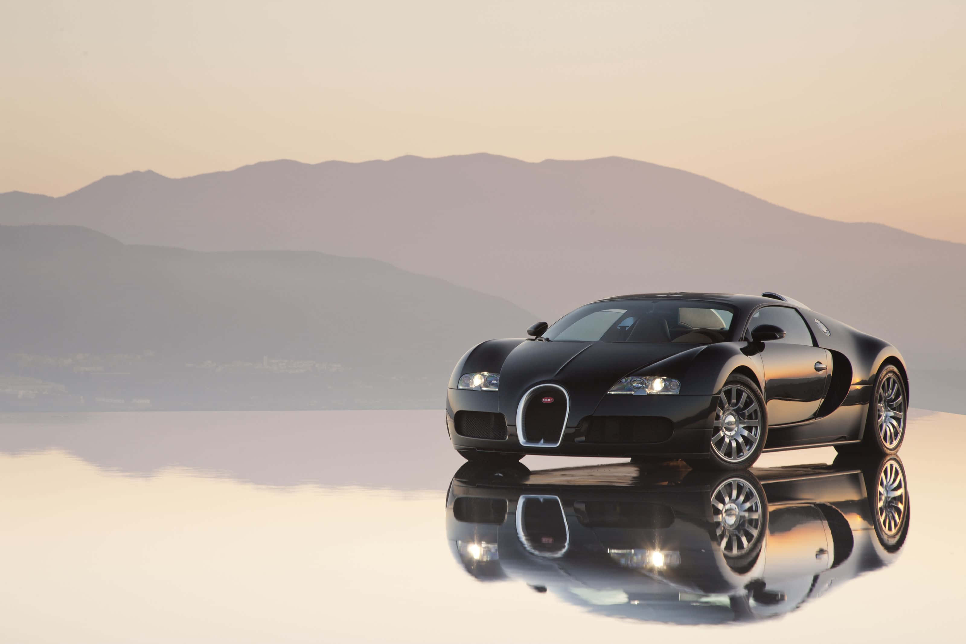 Bugatti Veyron 桌布 壁紙 — 一輛時尚的黑色布加迪超級跑車在反射表面上閃閃發光，薄霧籠罩的山脈在柔和的金色黎明光芒中。