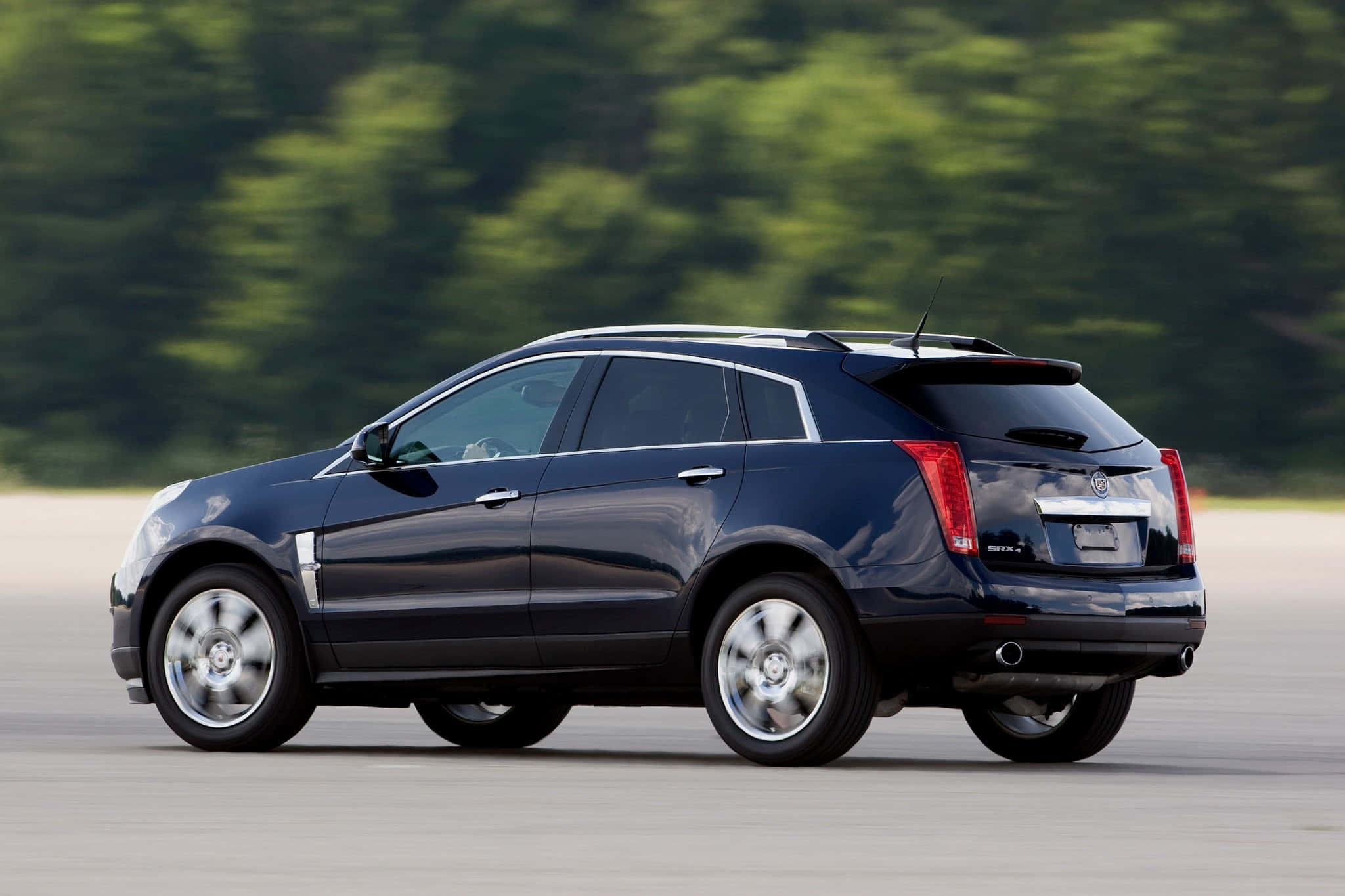 Cadillac Srx 桌布 壁紙 — 一輛時尚的黑色Cadillac SRX SUV在鋪砌的道路上行駛，背景有模糊的綠色樹木。