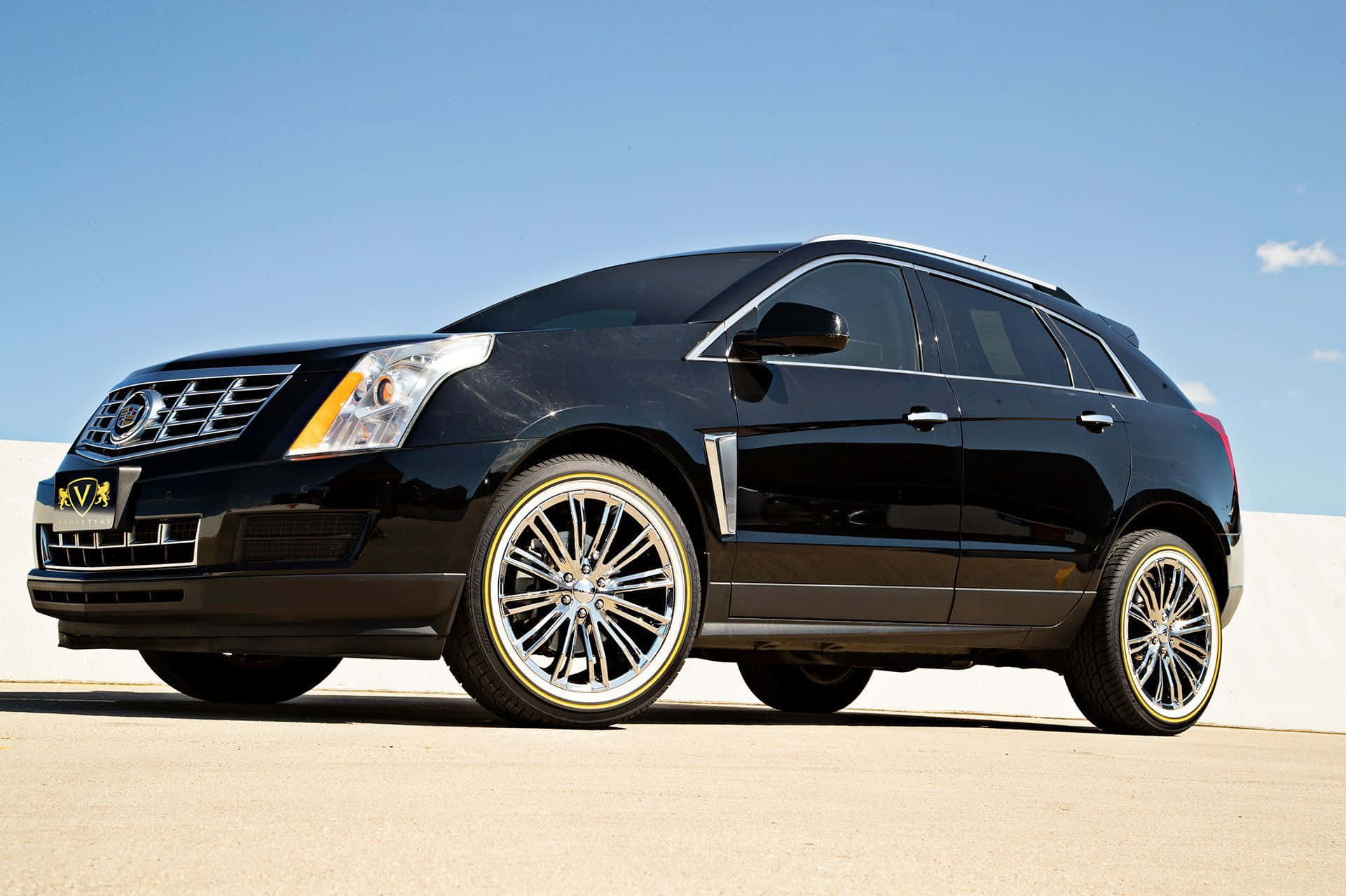 Cadillac Srx 桌布 壁紙 — 一輛時尚的黑色Cadillac SUV，配備定制金色輪圈，停泊在陽光普照的混凝土表面上，天空清澈湛藍。