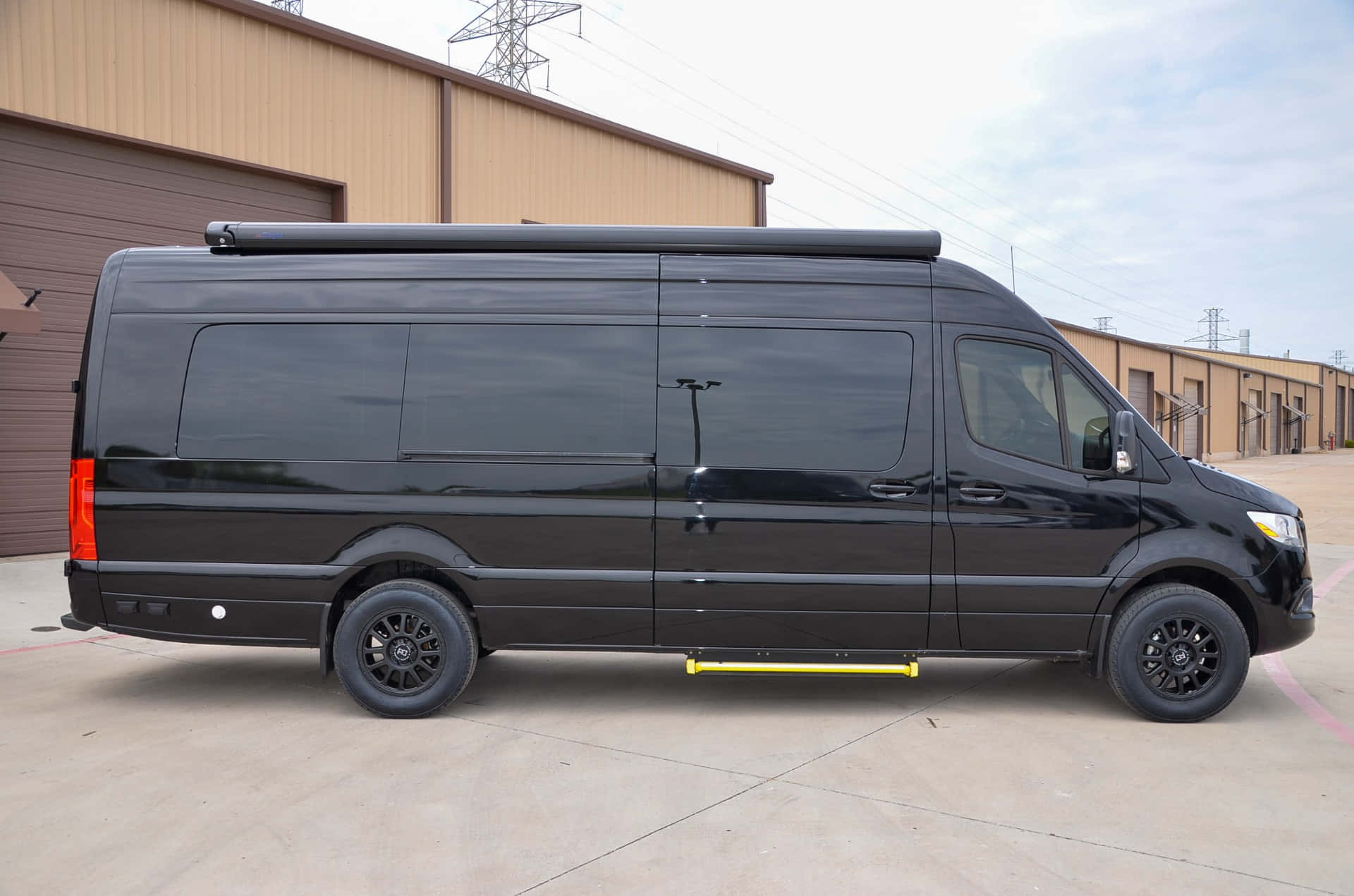 Mercedes Benz Sprinter 桌布 壁紙 — 一輛時尚的黑色露營車，配有車頂架和黃色踏板，停泊在工業建築前，天空多雲。