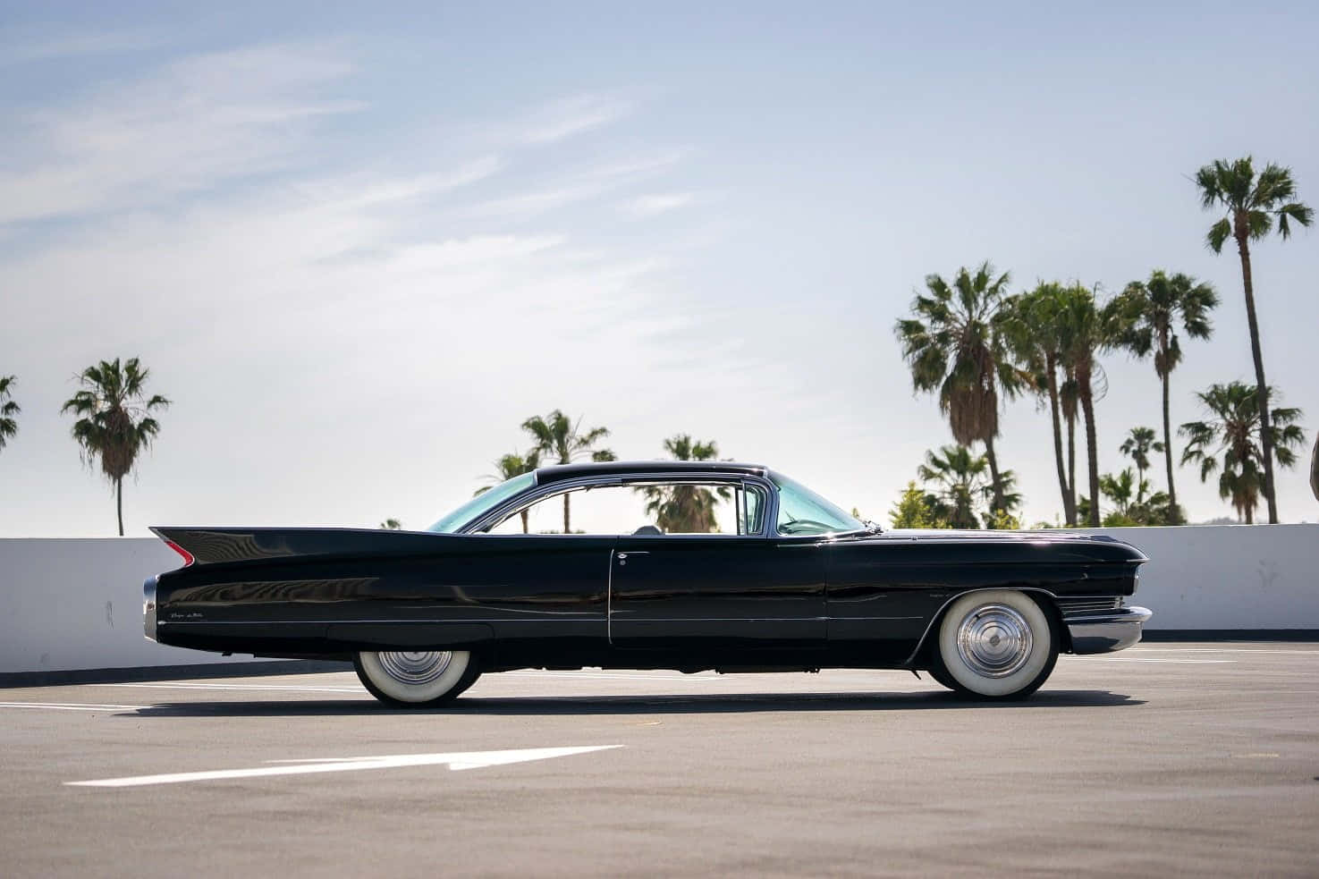 Cadillac Deville 桌布 壁紙 — 一輛時尚的黑色經典汽車，配備白色側壁輪胎，停放在陽光明媚的混凝土場地上，周圍環繞著高大的棕櫚樹。