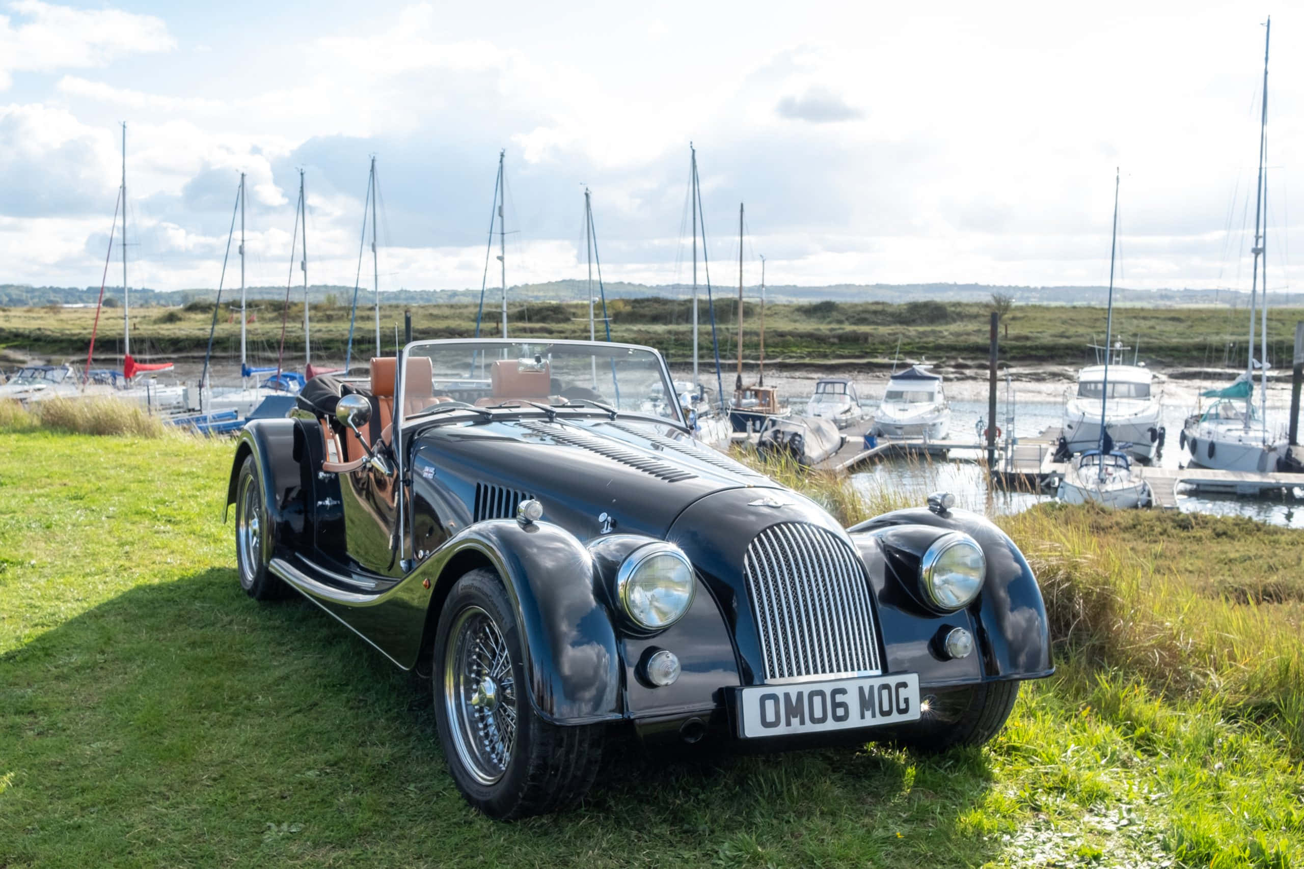 Morgan Roadster 桌布 壁紙 — 一架時尚的黑色經典開篷跑車停泊在草地上，旁邊是停滿帆船的碼頭，天空局部多雲。