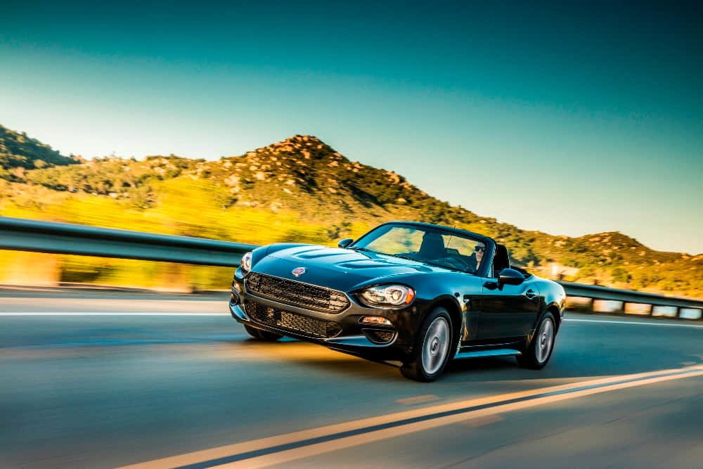 Fiat 124 Spider 桌布 壁紙 — 一架時尚的黑色敞篷跑車在晴朗藍天下沿著蜿蜒的山區高速公路飛馳。